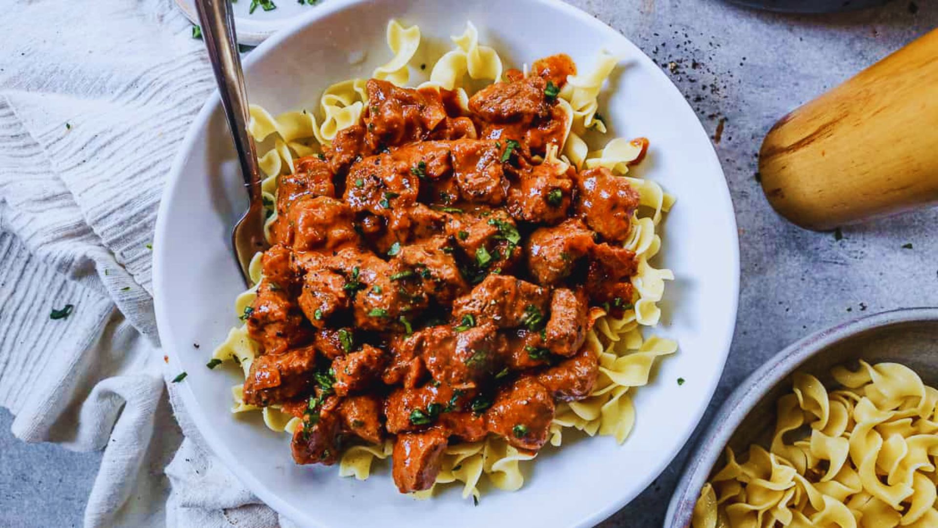 A bowl of beef stroganoff with noodles, garnished and accompanied by a silver spoon resting beside it.