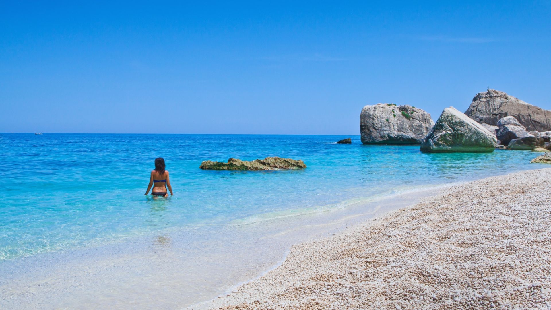 A woman stands waist-deep in the clear turquoise sea, facing large rocks and a bright blue sky. The sandy beach is in the foreground, evoking tranquility.