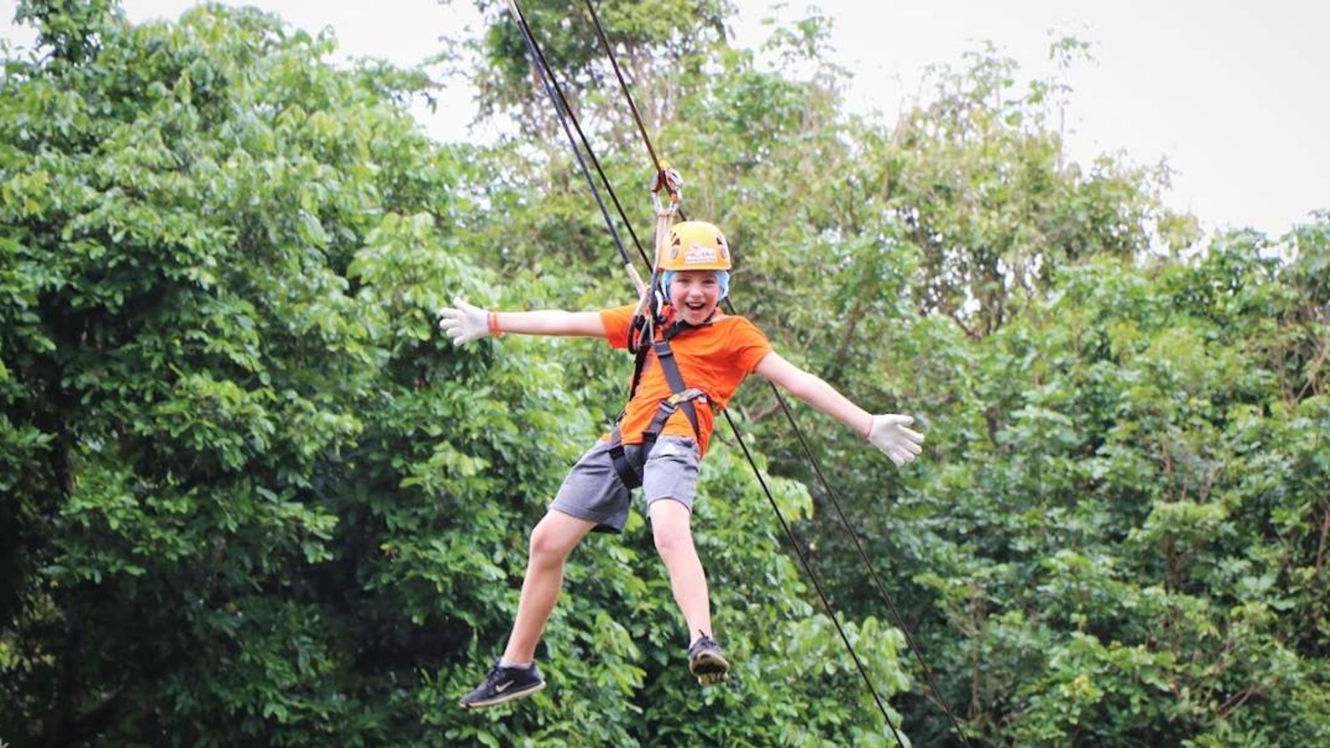 A boy in an orange shirt ziplining through trees, enjoying an adventurous outdoor experience.