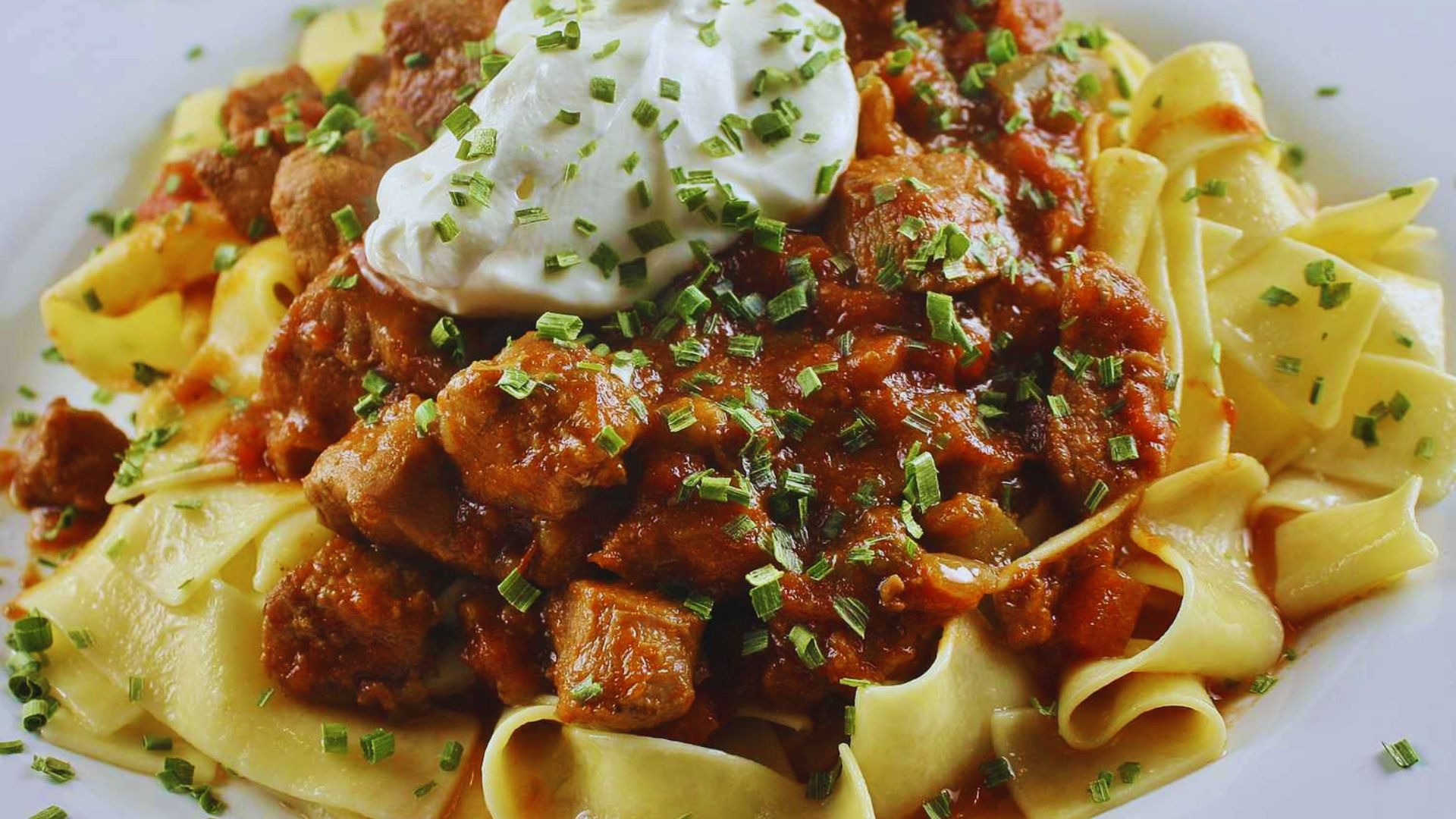 A plate of pasta topped with pork and drizzled with sour cream, garnished with herbs for added flavor.