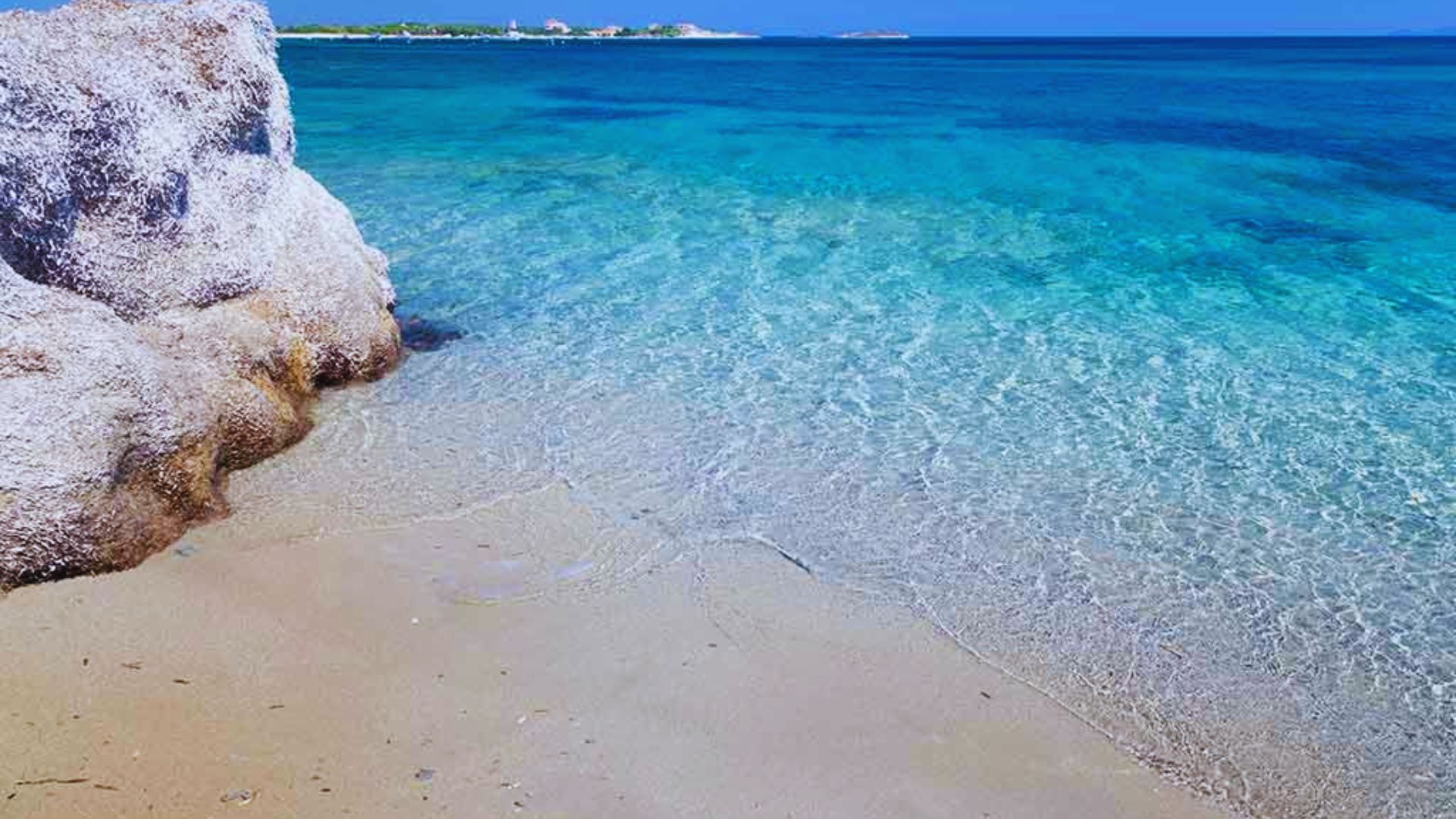 A tranquil beach scene with clear turquoise water gently lapping against a sandy shore, large rocks on the left, and a distant horizon under a bright blue sky.