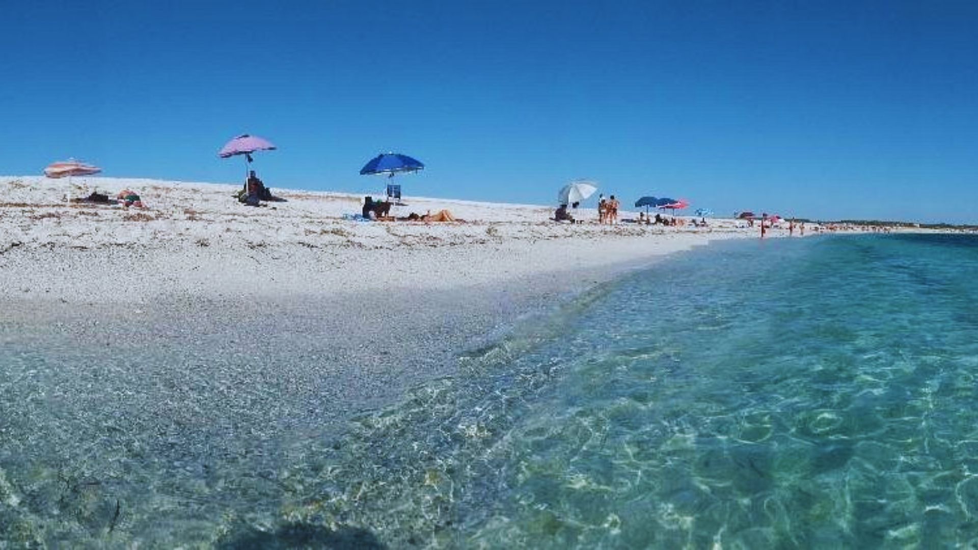 Crystal clear turquoise water laps against a sandy beach dotted with colorful umbrellas and sunbathers under a bright blue sky, conveying a serene, relaxing atmosphere.