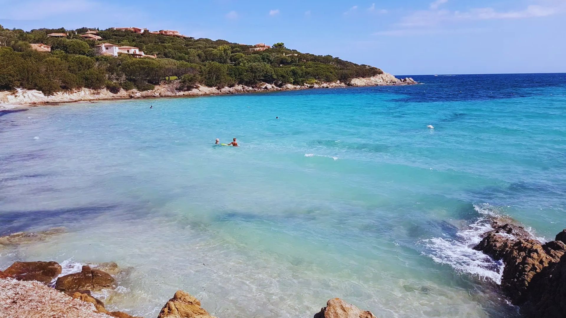 A serene beach with turquoise waters and gentle waves. People swim near the shore, backed by lush greenery and distant houses under a clear blue sky.