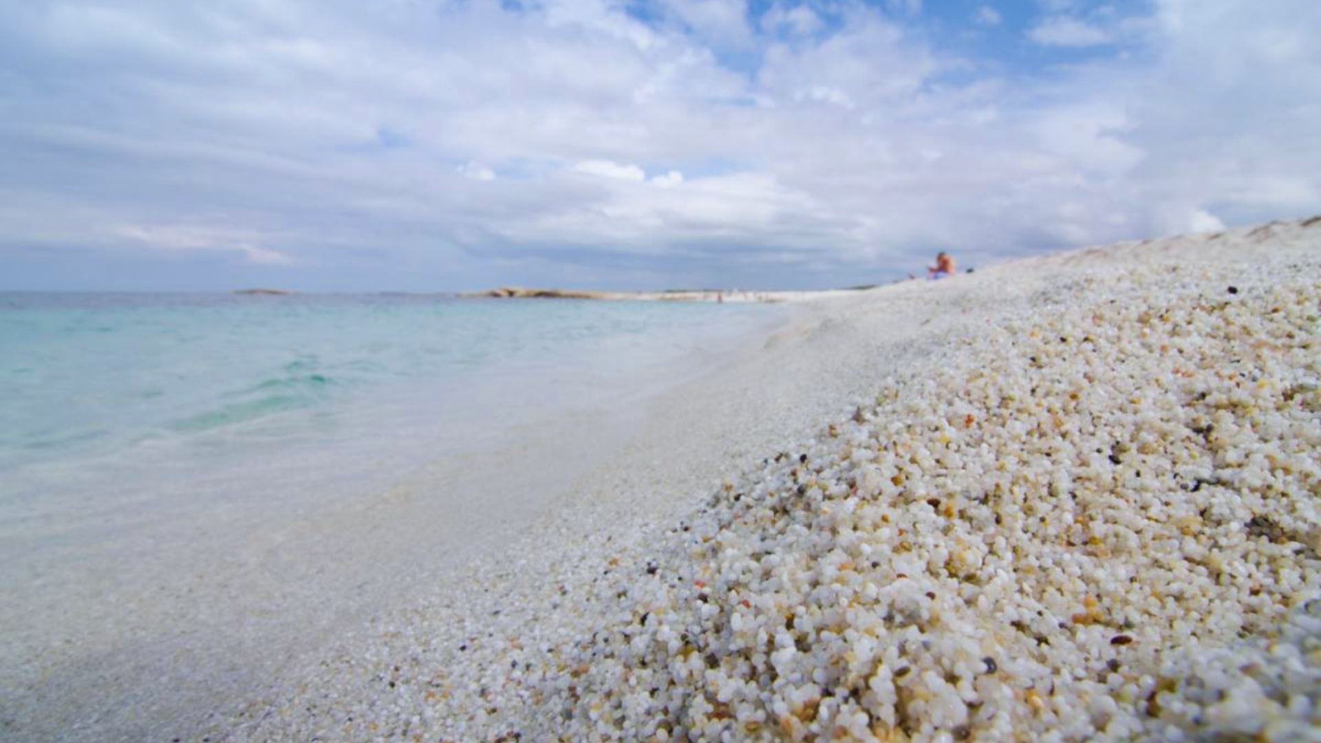 A serene beach scene with clear turquoise water meeting a shore covered in small, rounded pebbles. The sky is partly cloudy, creating a calm, peaceful atmosphere.