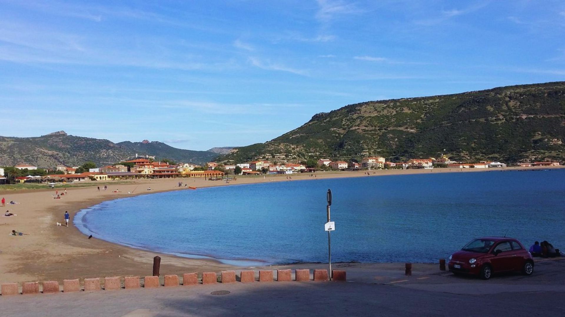 A serene coastal scene with a curving sandy beach and calm blue waters, bordered by hills. People relax on the sand, and a car is parked nearby.