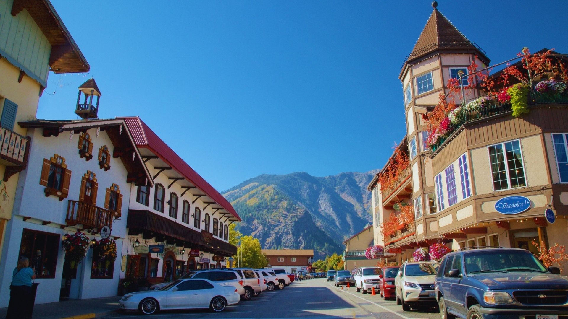 A mountain town street lined with parked cars, surrounded by scenic mountain views.