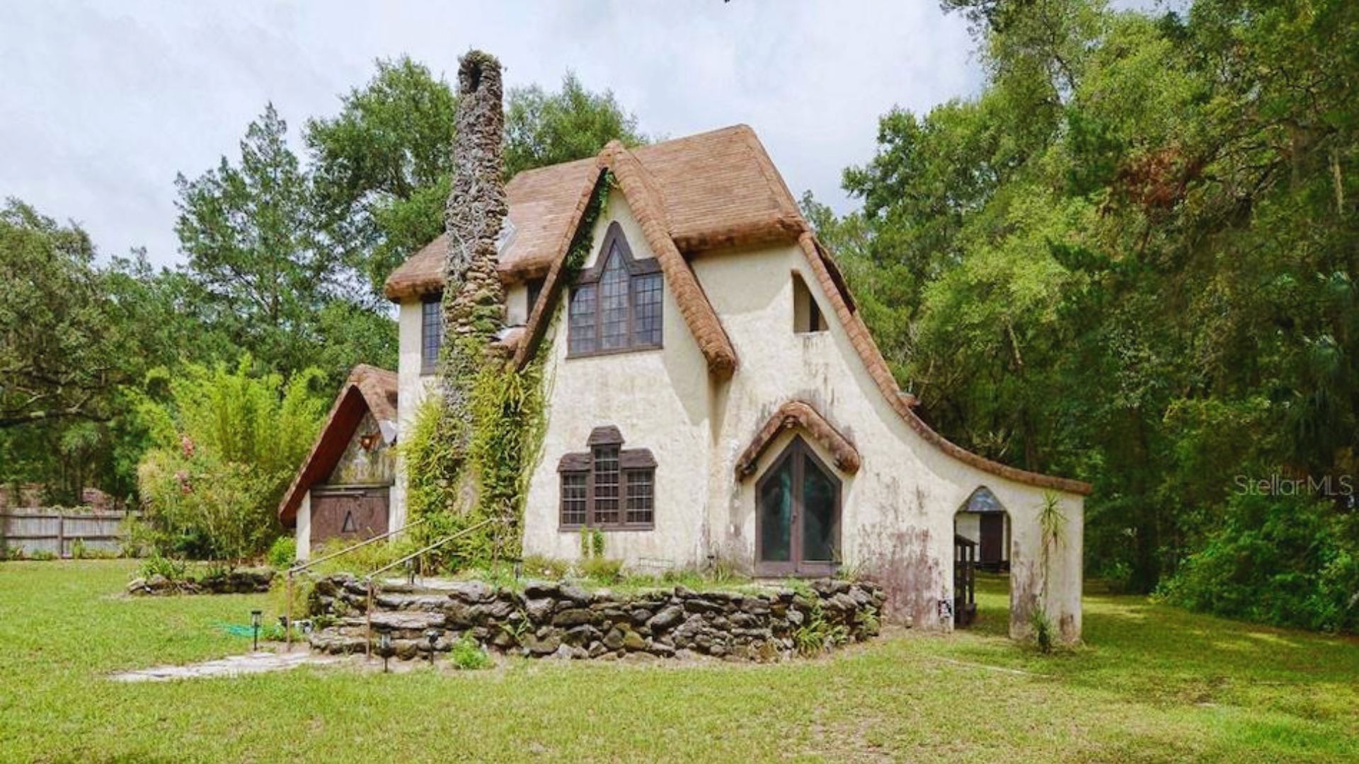 A whimsical cottage with a steep, thatched roof and stone chimney is surrounded by lush greenery, exuding a fairytale-like charm and peacefulness.