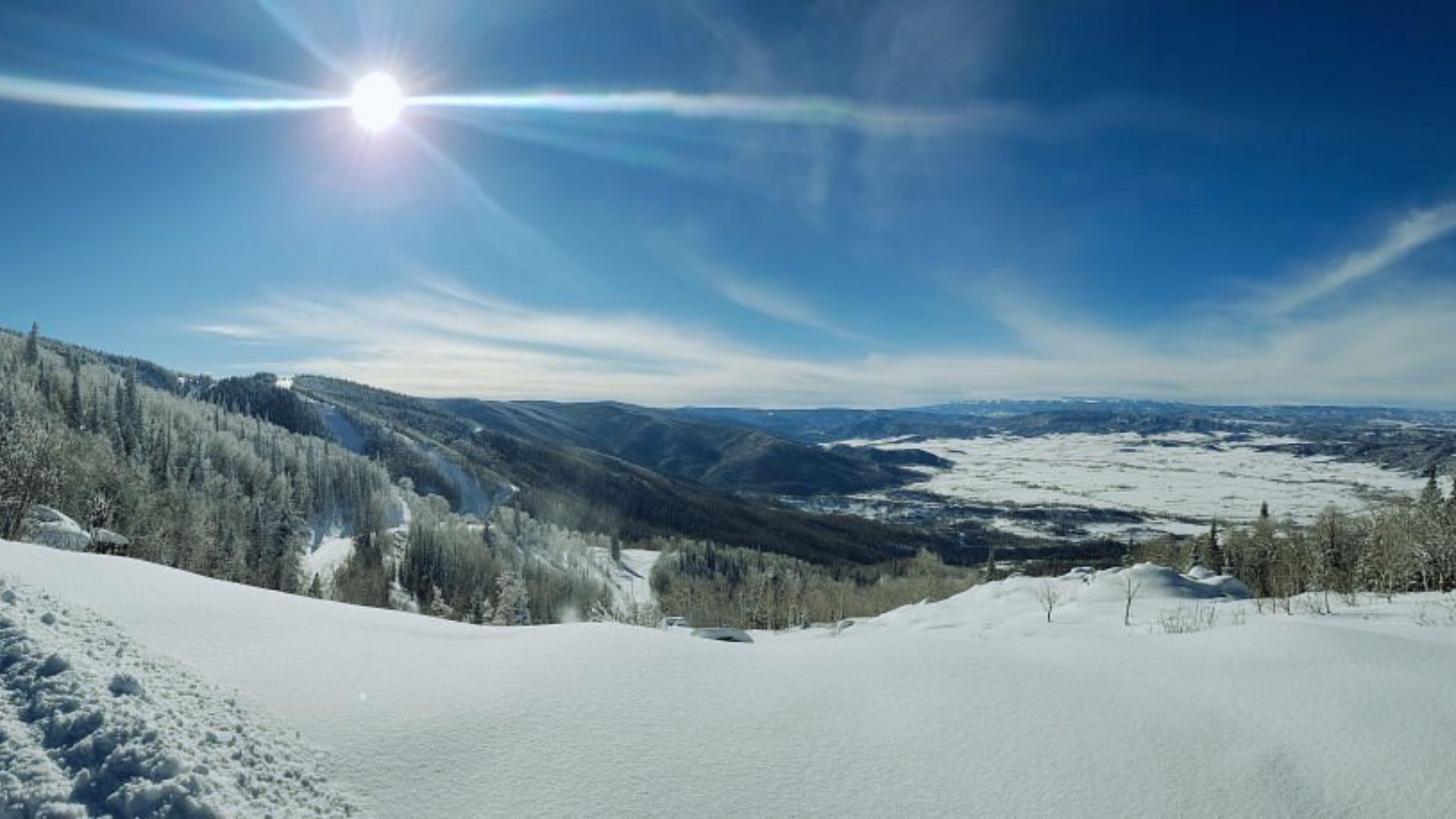 Snowy landscape with snow-covered hills and dense pine forests under a bright, radiant sun. Clear blue sky enhances the serene, crisp winter atmosphere.