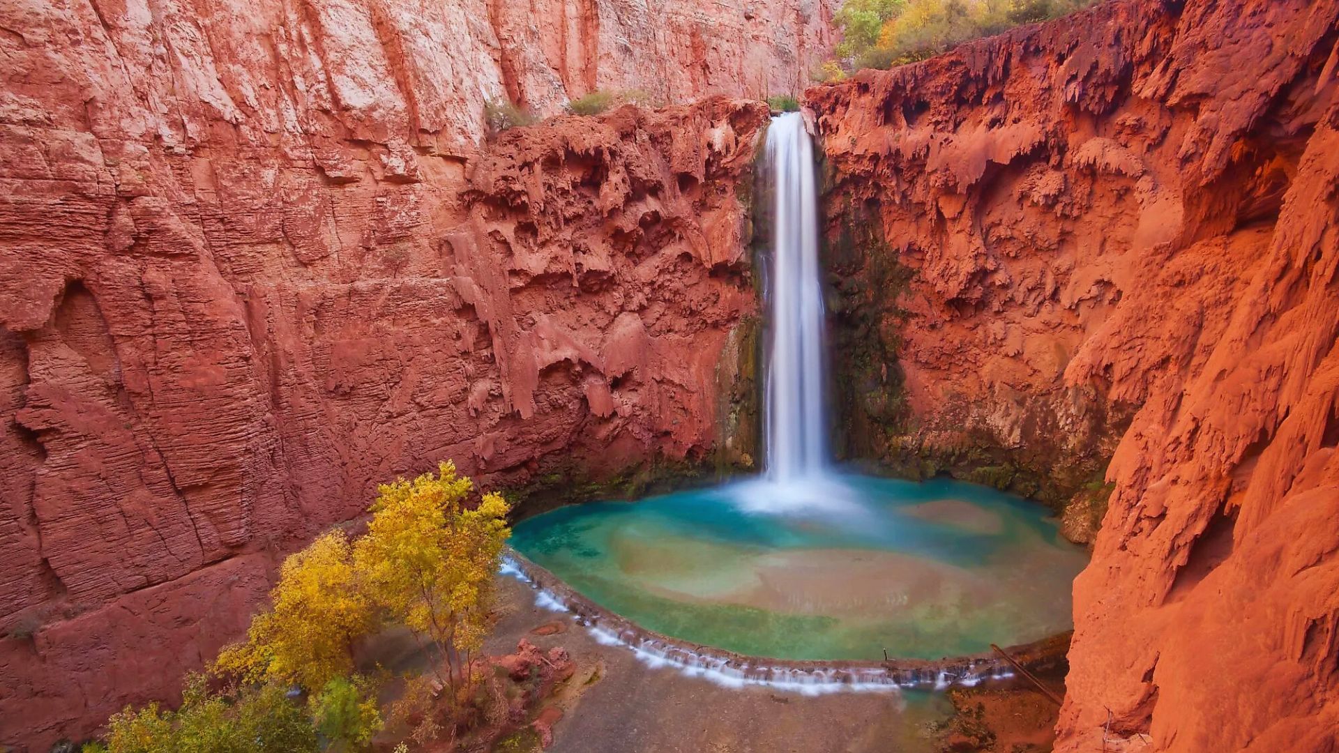 A vibrant waterfall cascades into a turquoise pool, surrounded by striking red rock cliffs. Autumn trees add a touch of gold. Serene and majestic.