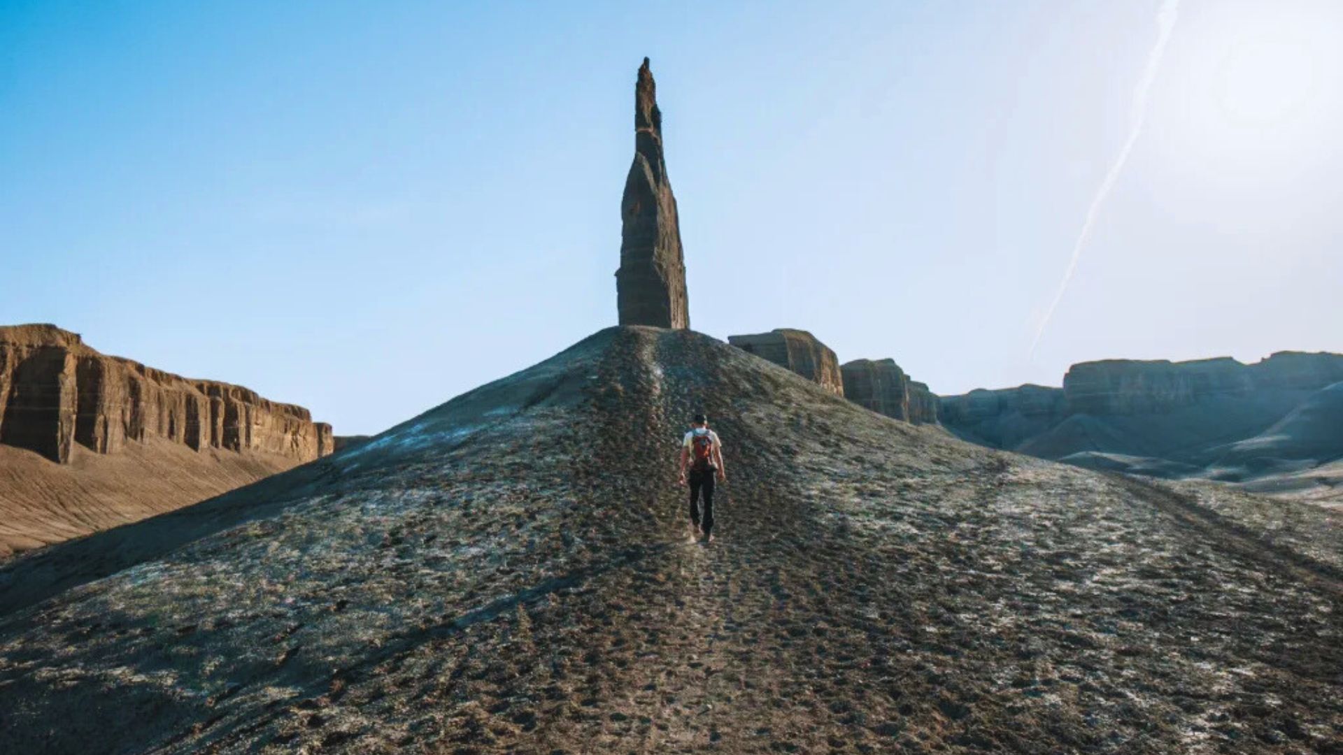 Person in outdoor gear walking up a rocky hill toward a tall, narrow rock formation under a clear blue sky. The scene conveys a sense of adventure and isolation.