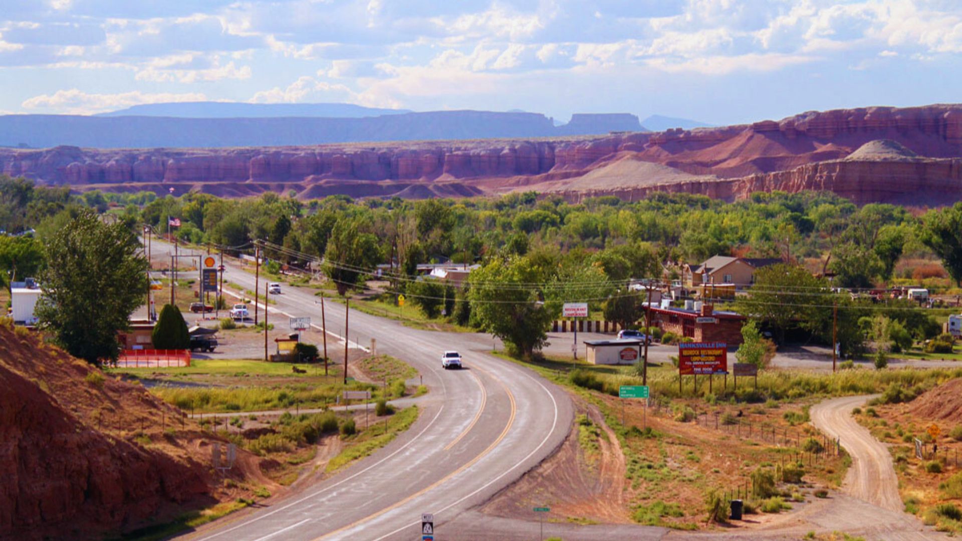 A scenic road curves through a small town, surrounded by lush greenery and red rock formations under a partly cloudy sky, conveying a serene atmosphere.