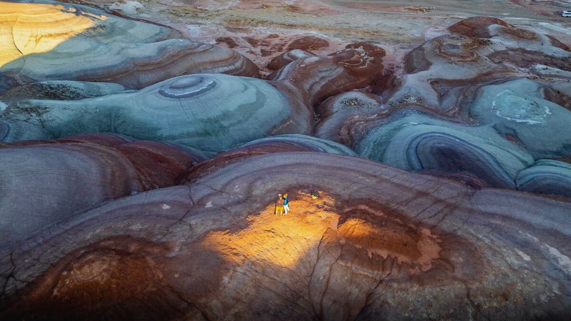 Aerial view of a person in bright clothes standing on colorful, swirling rock formations with red, brown, and blue hues, creating a surreal, dreamlike scene.