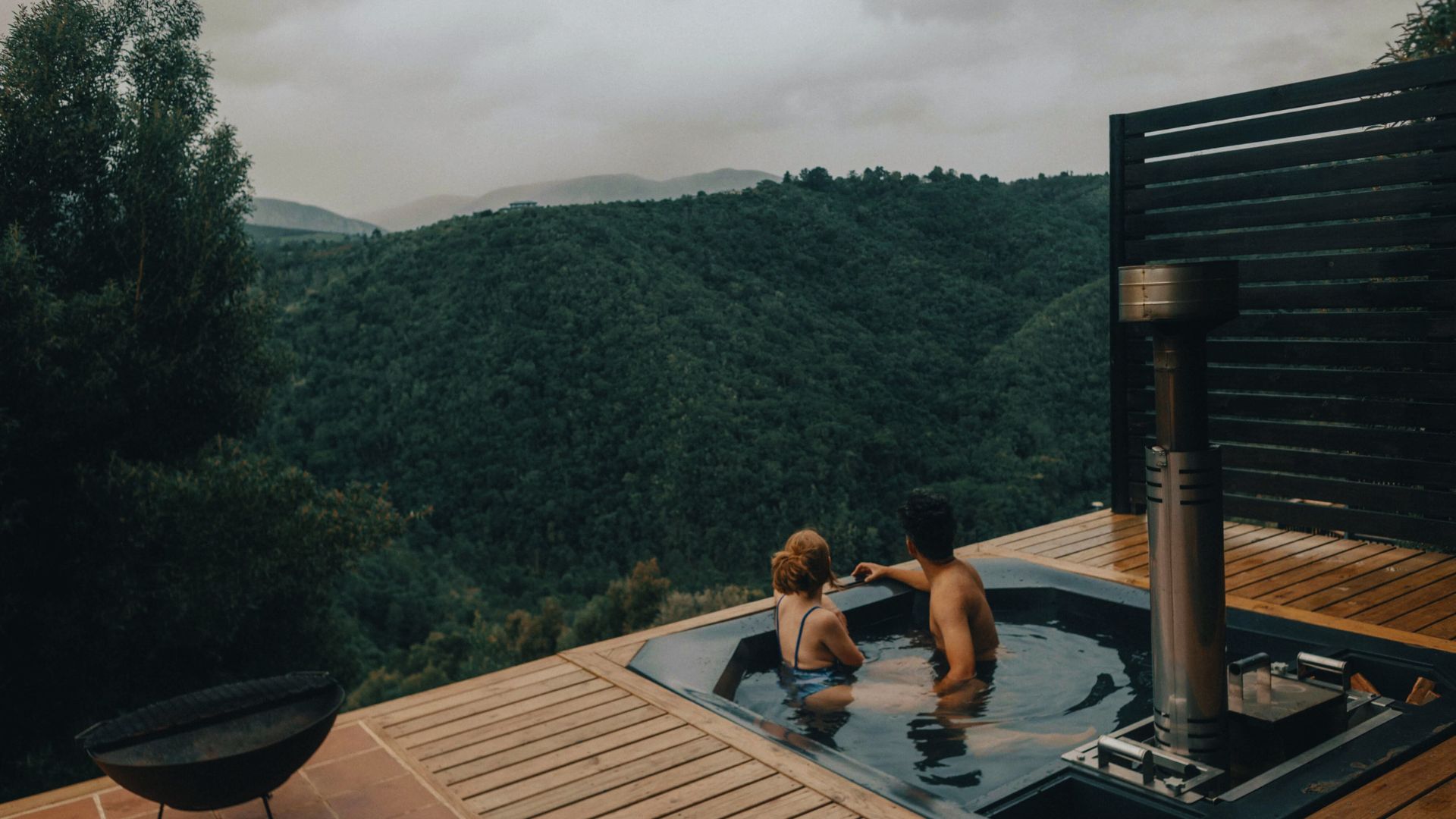 Two people relax in a hot tub on a deck, enjoying a scenic view of the mountains in the background.