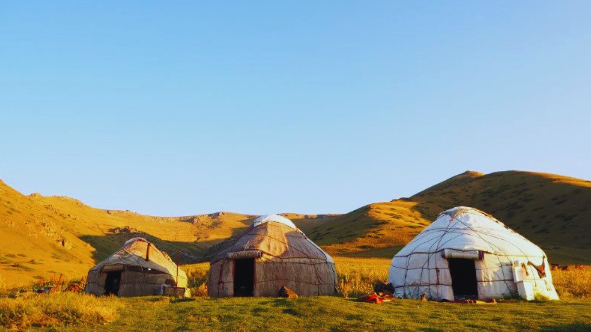 Three rustic huts nestled in the mountains, surrounded by lush grass and towering peaks in the background.