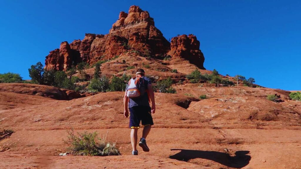 Bell Rock Vortex Hike Guide & Trail Info A man carrying a backpack walks along a scenic trail surrounded by trees and greenery.