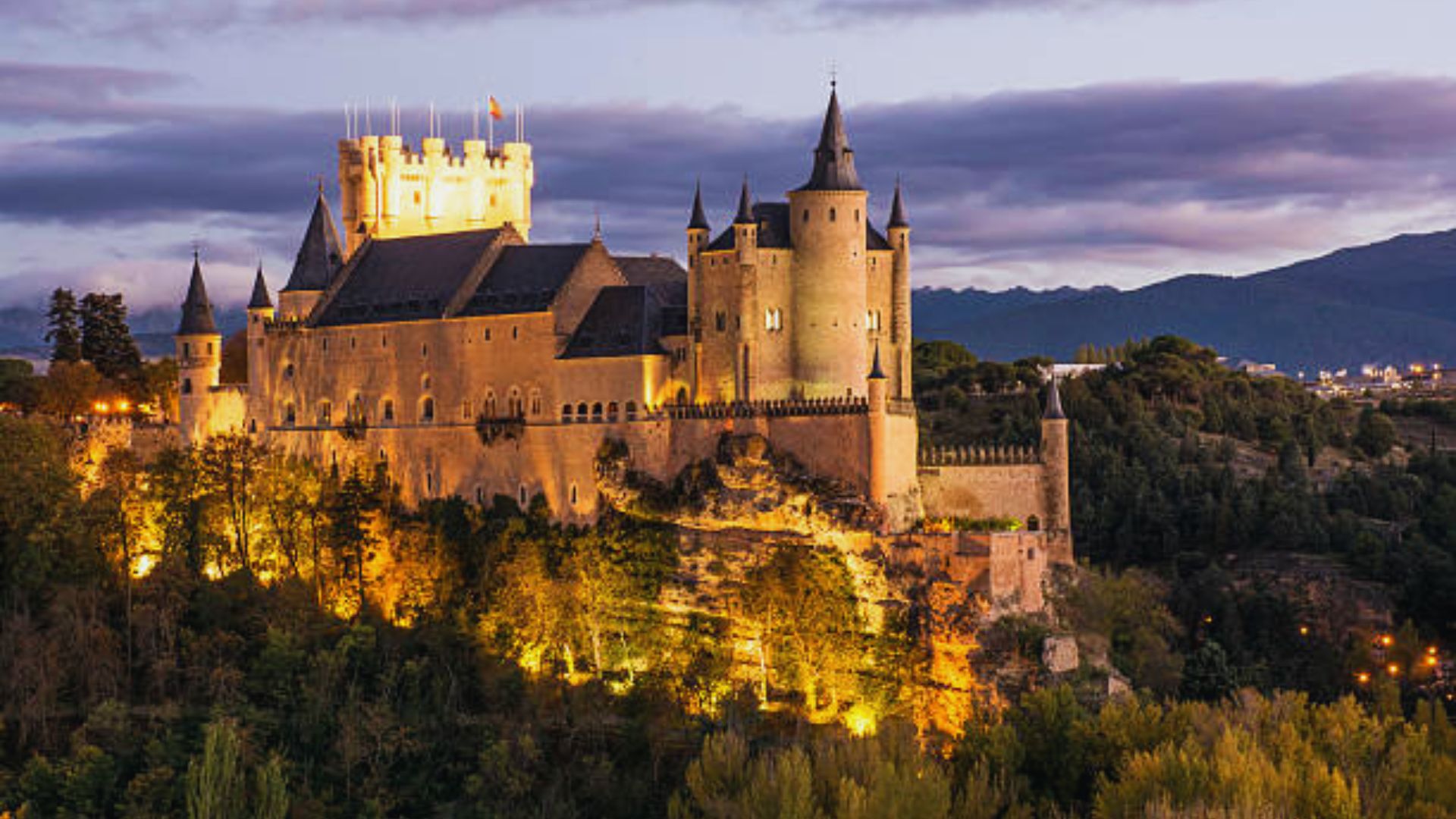 Majestic castle illuminated at dusk, perched on a hill with gothic spires and turrets. Clouds fill the sky, creating a dramatic and enchanting atmosphere.