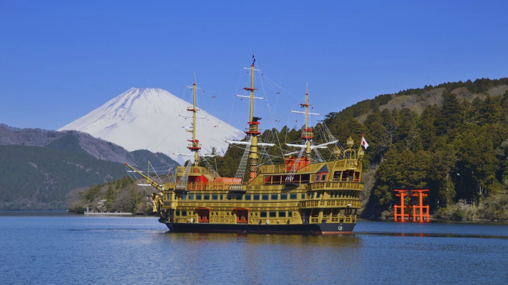 A golden pirate ship sails on a calm lake with Mount Fuji in the background. A red torii gate stands on the right, surrounded by lush green trees under a clear blue sky.