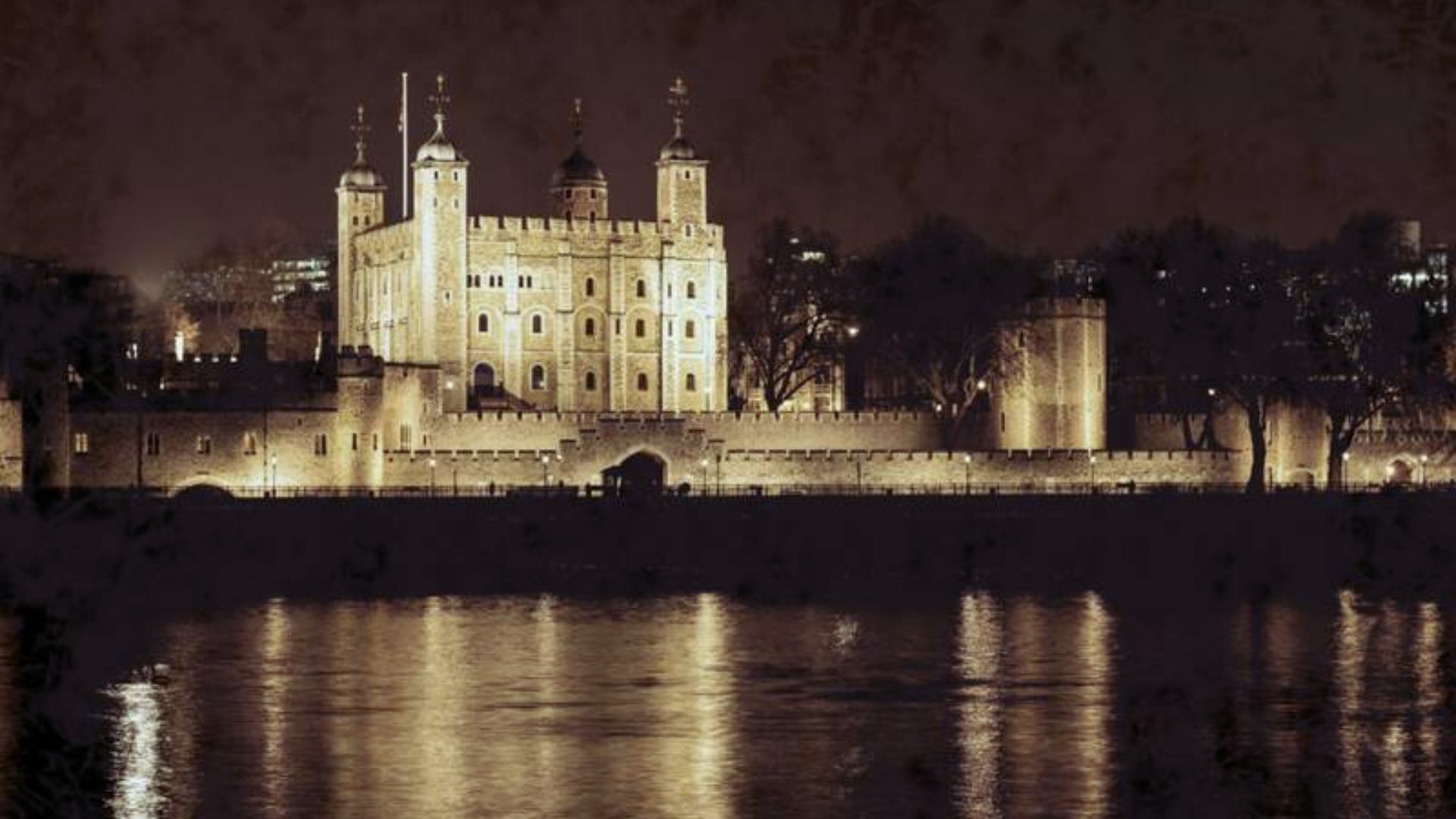 A well-lit castle with four towers stands majestically at night, its reflection shimmering on a calm river, creating a serene and historic atmosphere.