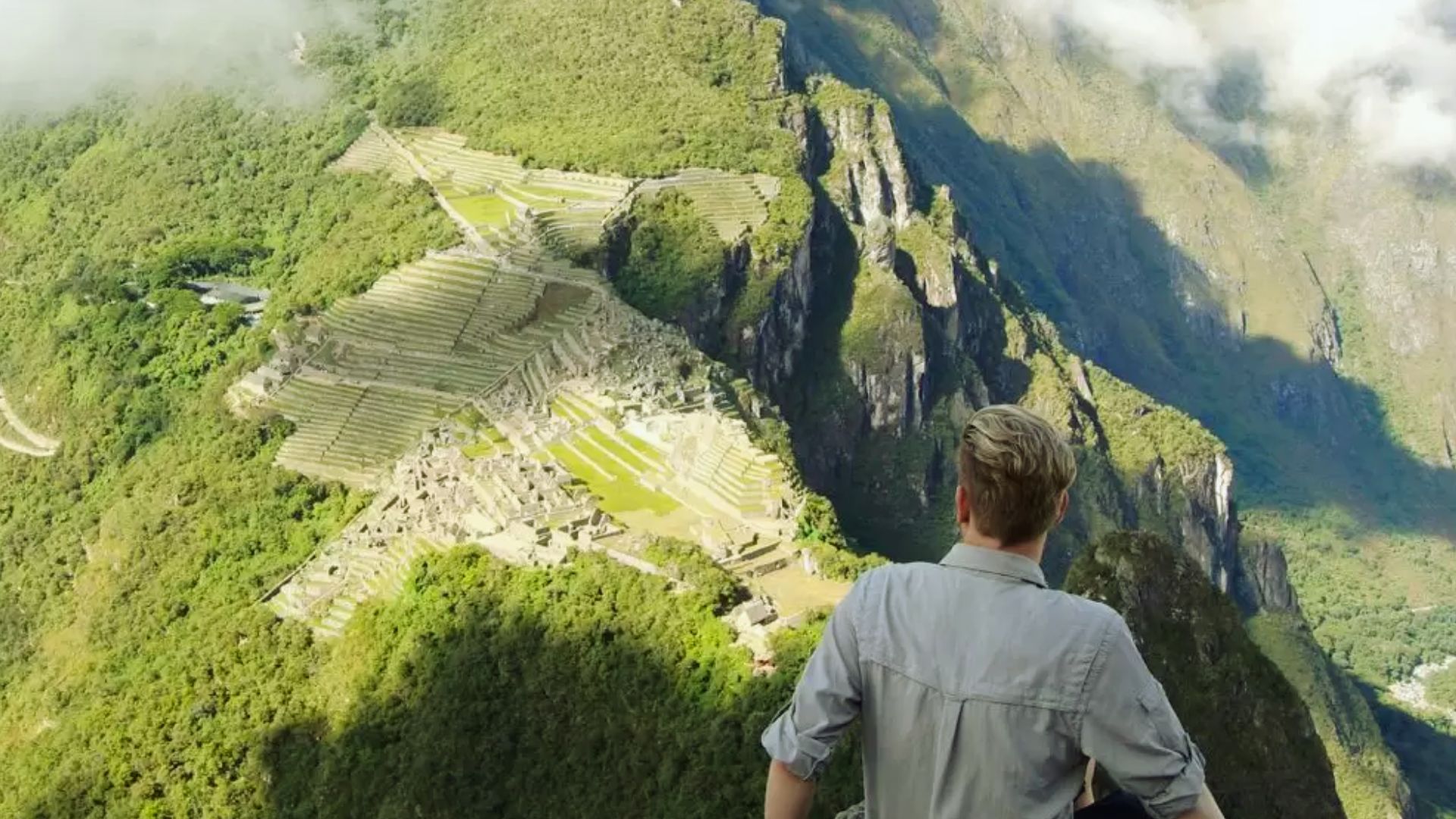 A person with short hair stands on a cliff, looking at the ancient ruins of Machu Picchu. The lush green mountains surround the site under a cloudy sky.
