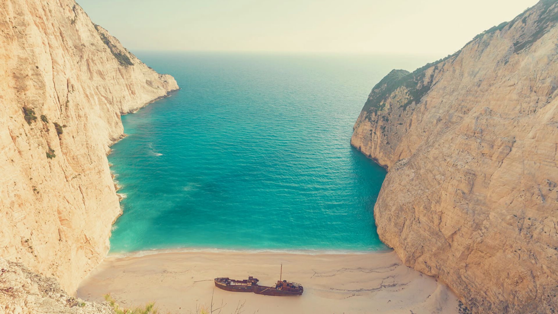 A serene cove with turquoise water flanked by steep cliffs. A rusted shipwreck rests on the sandy beach, creating a scene of tranquil isolation.