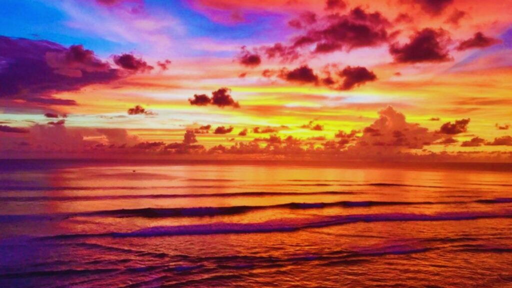 Vibrant sunset over the ocean with fiery hues of red, orange, and purple in the sky, reflecting on gently rippling waves below. Tranquil and breathtaking.