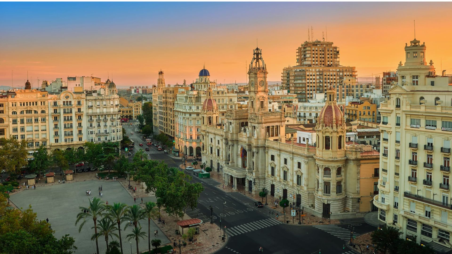 Aerial view of a historic square at sunset, featuring ornate buildings with domes and towers. The glowing sky adds a warm, serene atmosphere.