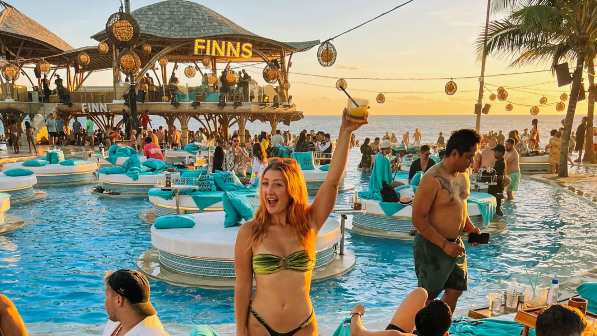 A lively beach club scene at sunset with people enjoying a pool. A woman in a green bikini raises a cocktail, surrounded by turquoise lounge chairs and palm trees.