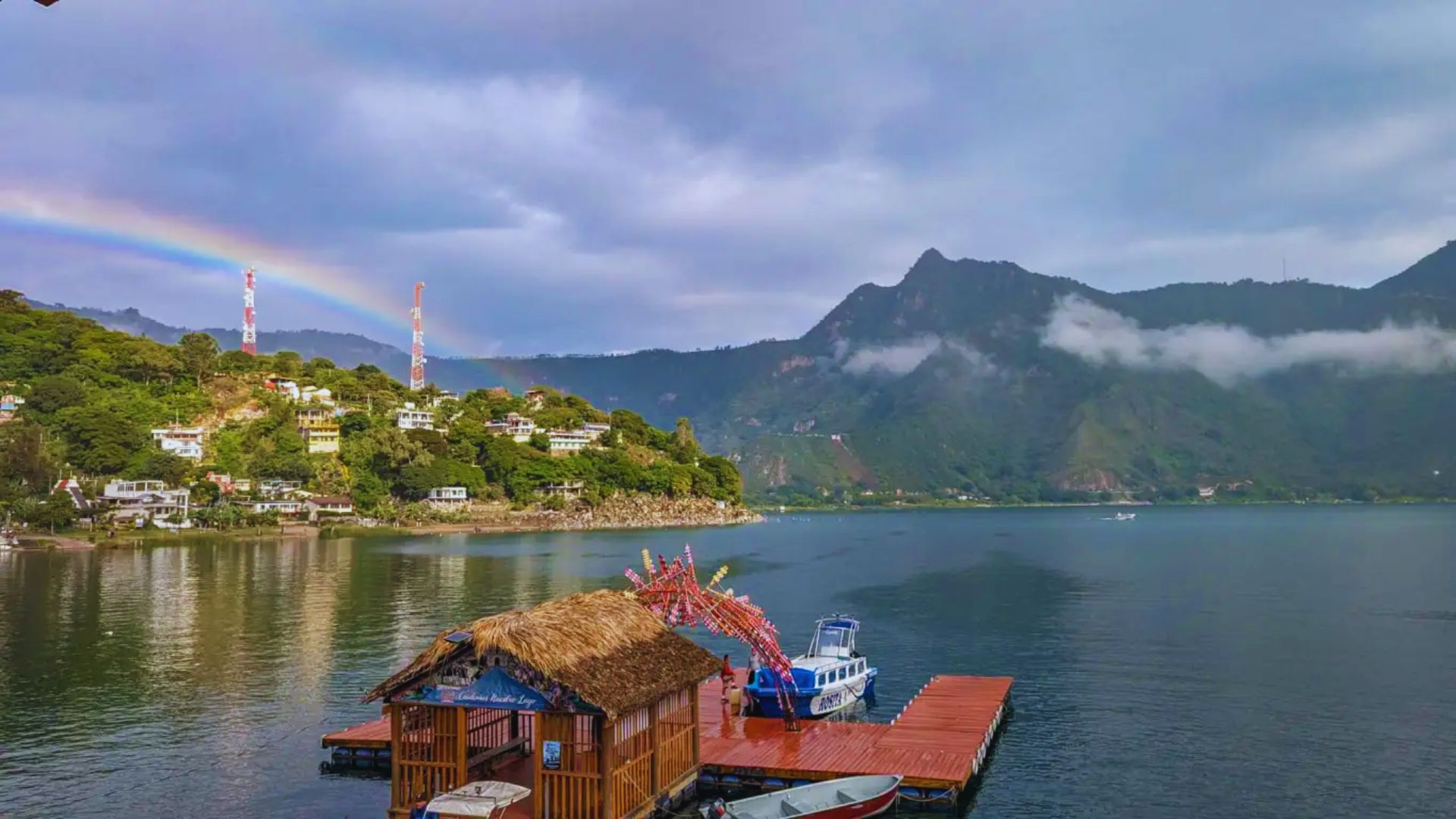 A serene lakeside scene with a wooden dock and thatched hut, surrounded by lush hills. A rainbow arcs over the village, evoking a peaceful, idyllic vibe.