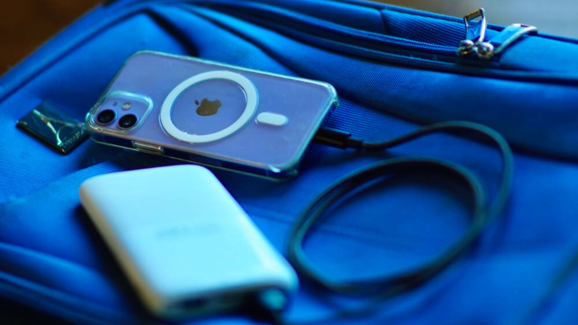 A smartphone with a transparent case and a charging cable lies on a vibrant blue bag, next to a square power bank. The scene conveys readiness and connectivity.