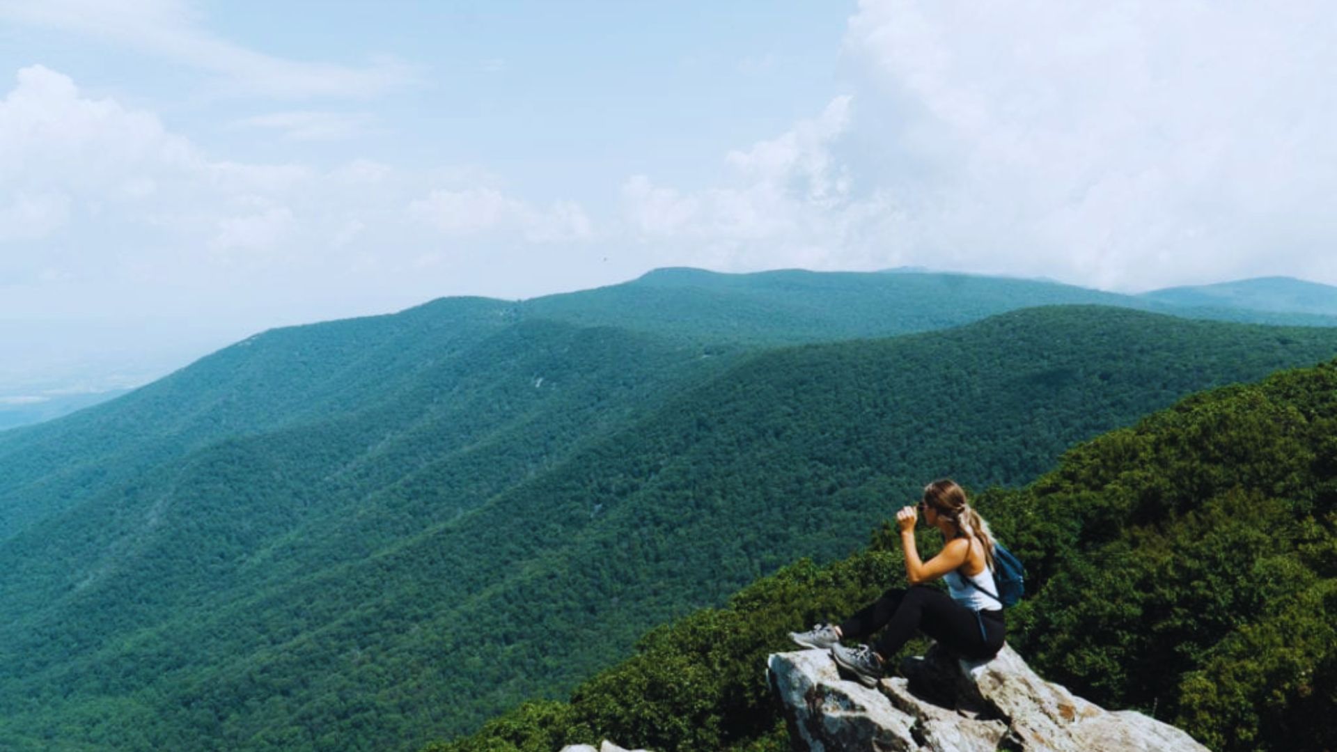 A person sits on a rocky outcrop, taking photos of expansive, lush green mountains under a blue sky, conveying a sense of adventure and tranquility.