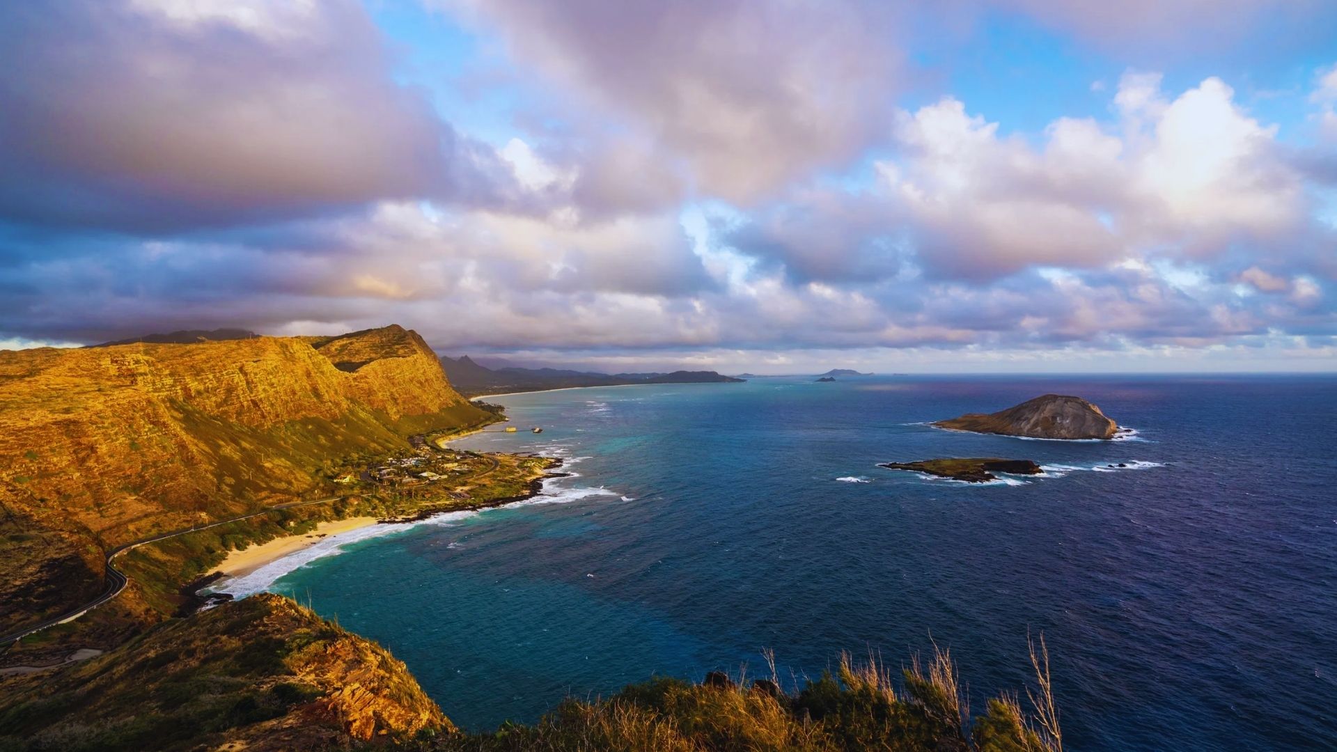 Vibrant coastal landscape with a golden cliff on the left, a sandy beach below, and blue ocean extending to the horizon under a cloudy sky.