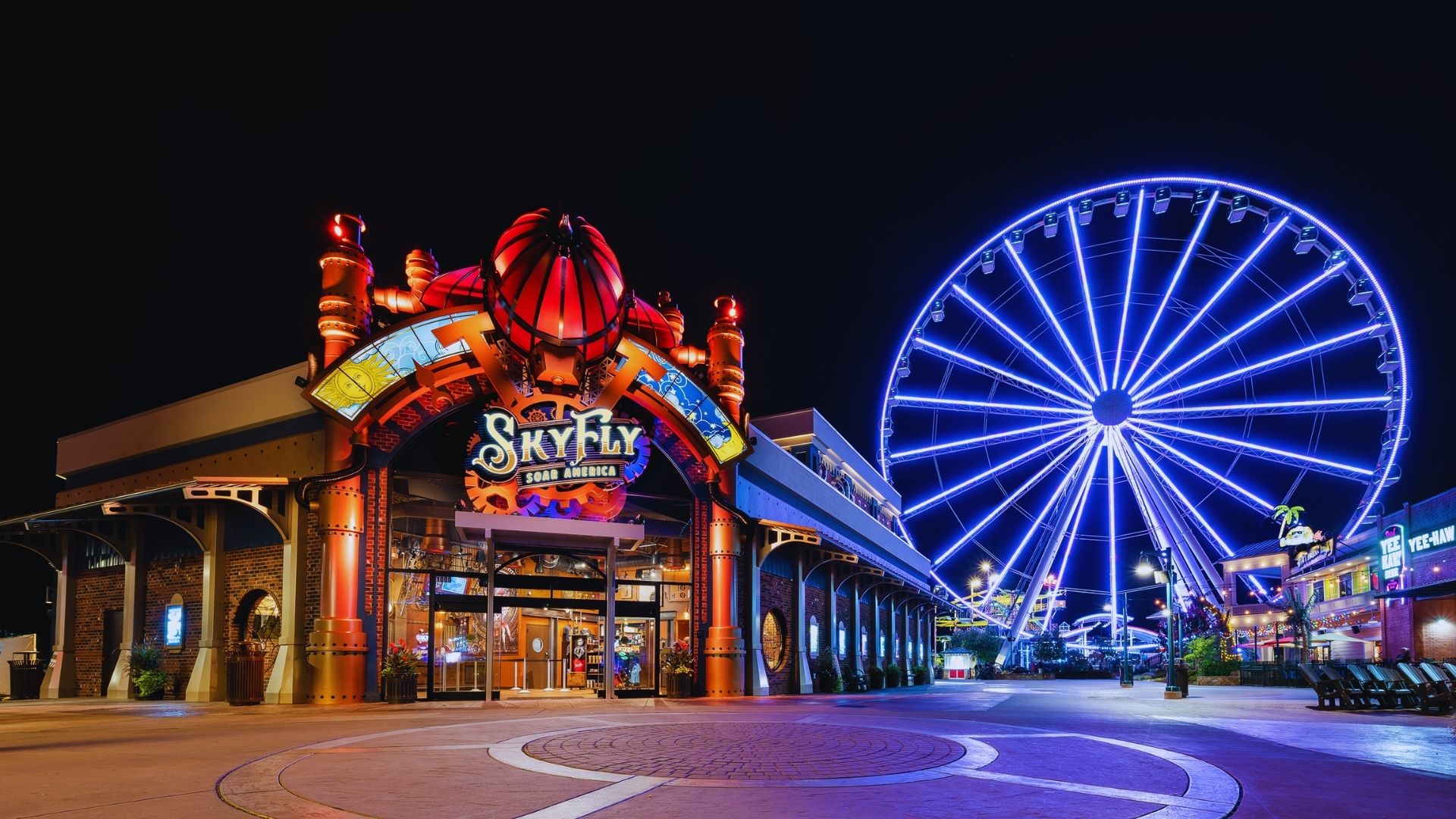Brightly lit amusement park at night with SkyFly attraction entrance on the left and a large illuminated Ferris wheel on the right, creating a lively atmosphere.