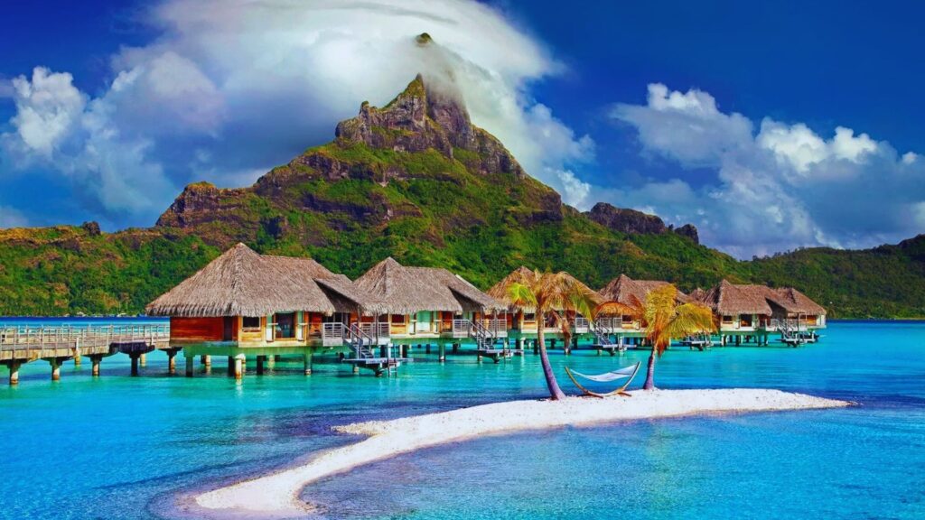 13 Best Caribbean Islands to Visit for Family Holidays Tropical paradise with overwater bungalows, a small sandy island with palm trees and a hammock, turquoise water, and a lush mountain under a cloudy sky.