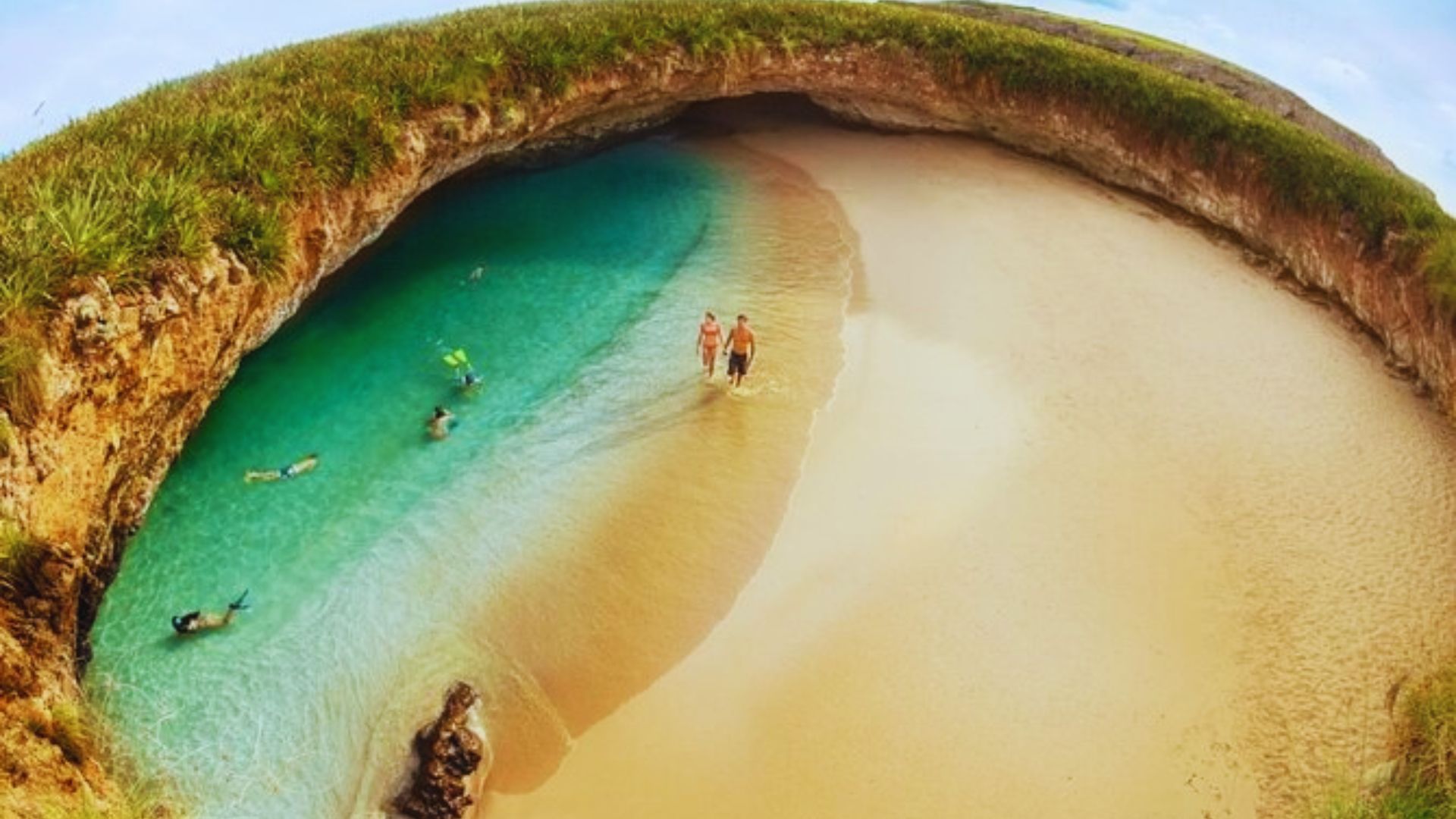 Hidden beach with turquoise water and golden sand, surrounded by rocky cliffs with green vegetation. People swim and walk, creating a serene atmosphere.