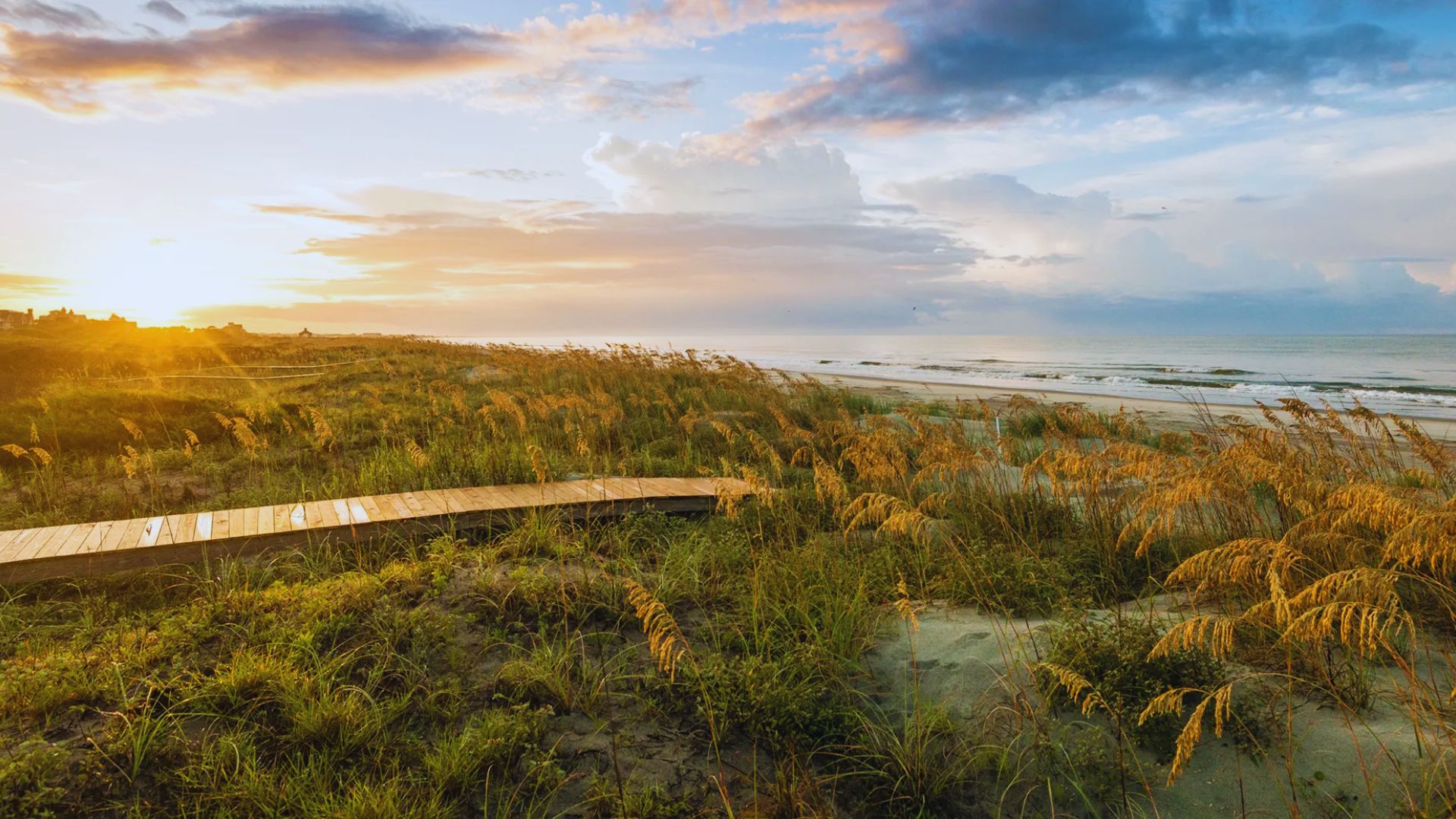 Sunset view over a serene beach, with golden light illuminating tall grasses and a wooden path leading to the sandy shore. The sky is vibrant with clouds.