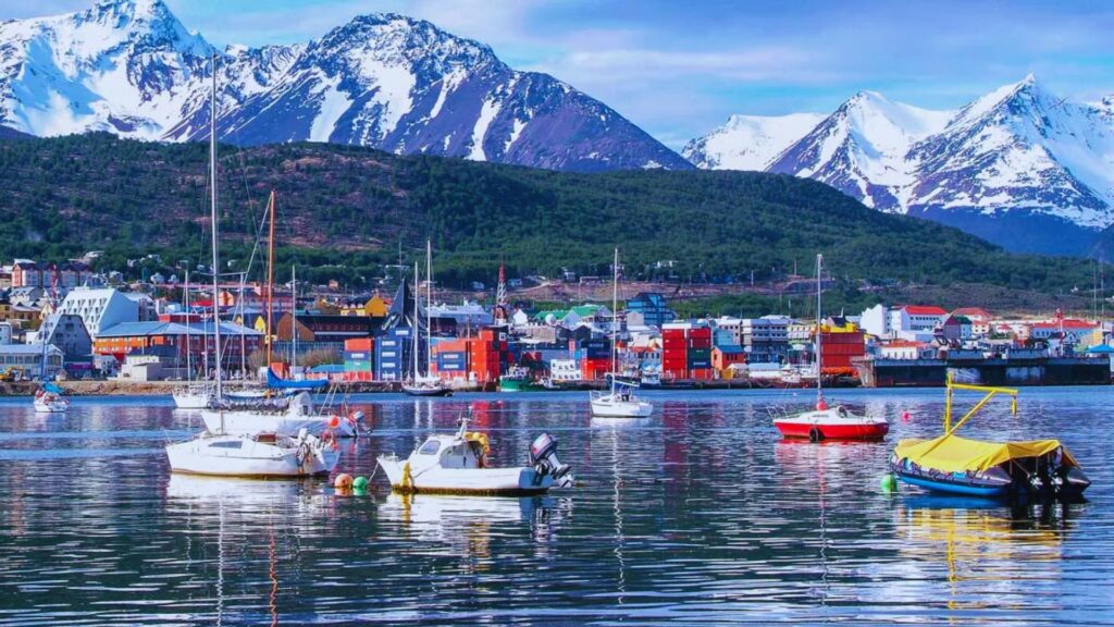 Colorful boats float on a calm harbor with vibrant buildings and snow-capped mountains in the background, creating a serene and picturesque scene.