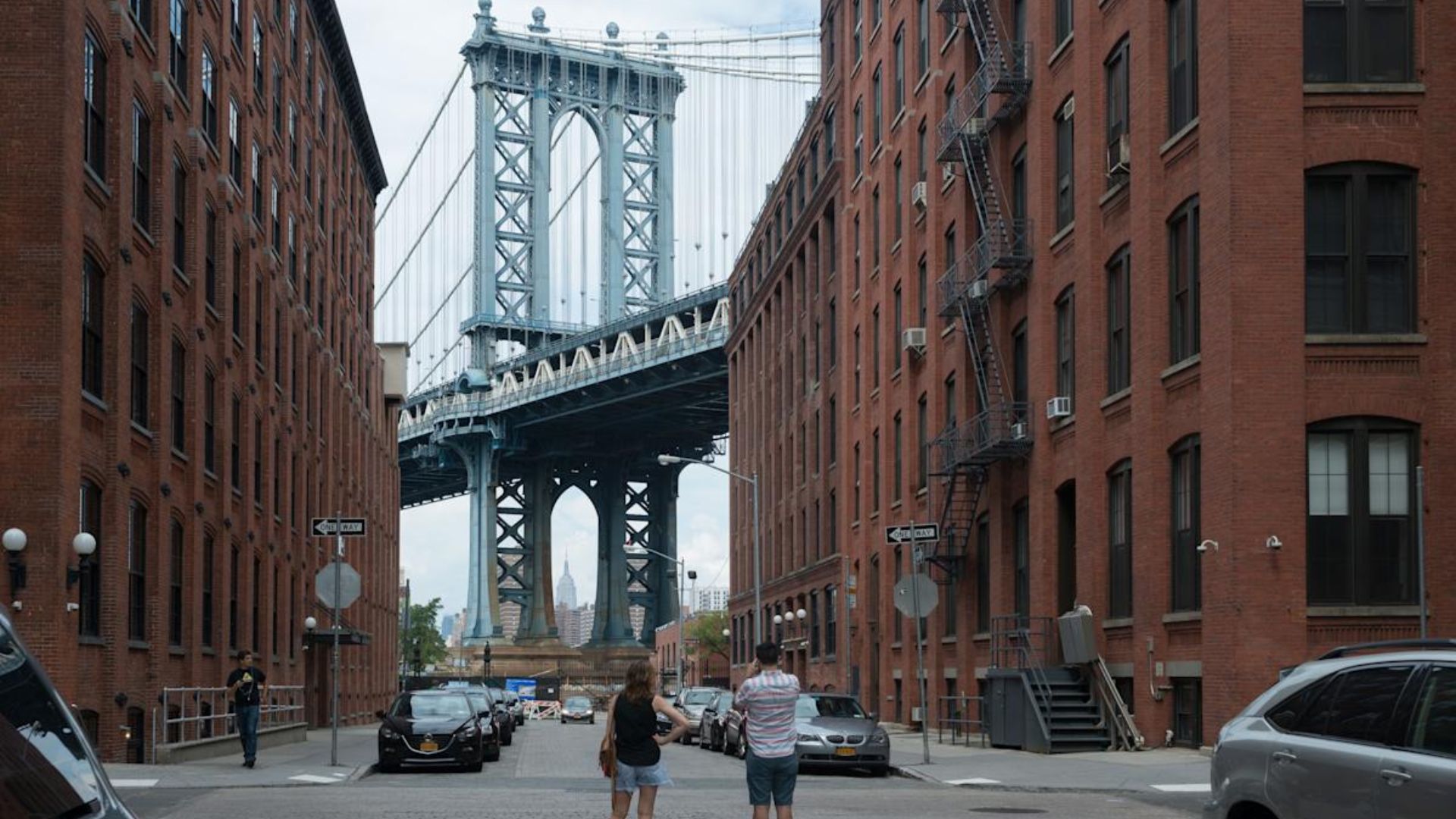 A pair of individuals strolls down an alley, with a bridge seen in the distance.