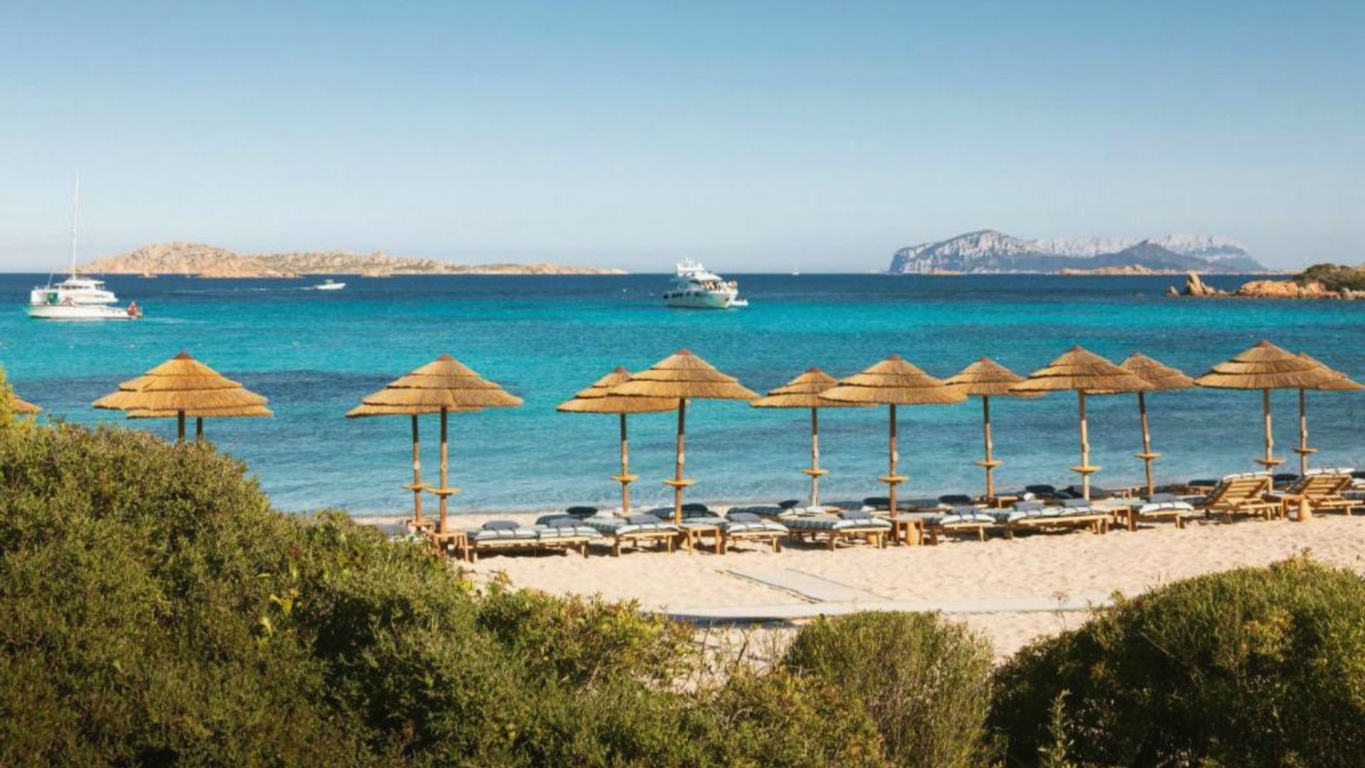 Sandy beach with rows of thatched umbrellas and lounge chairs, overlooking turquoise sea and distant islands. Boats float on the calm water, under a clear blue sky.