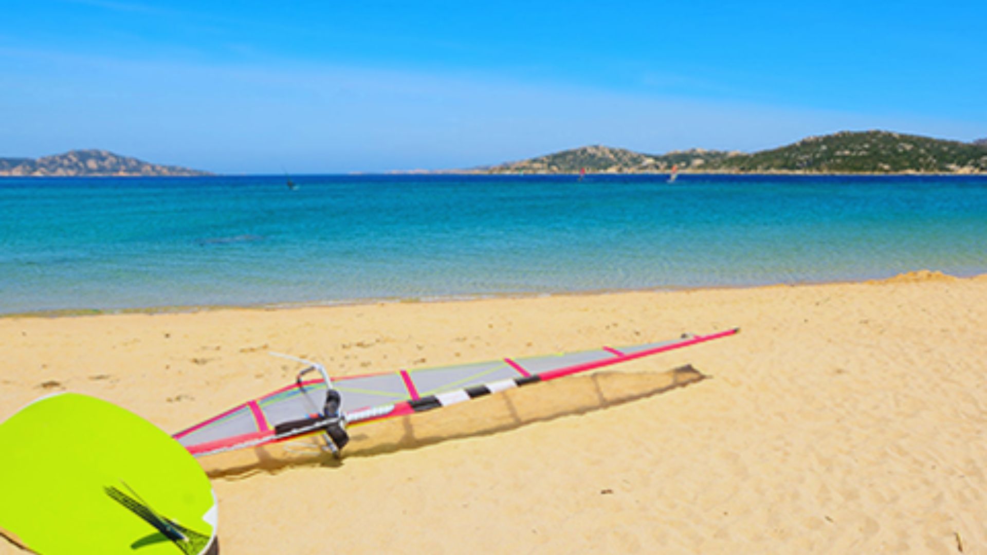 A serene beach scene with a vibrant blue ocean and clear sky. A windsurf board and sail are resting on the golden sand, evoking a sense of relaxation and leisure.