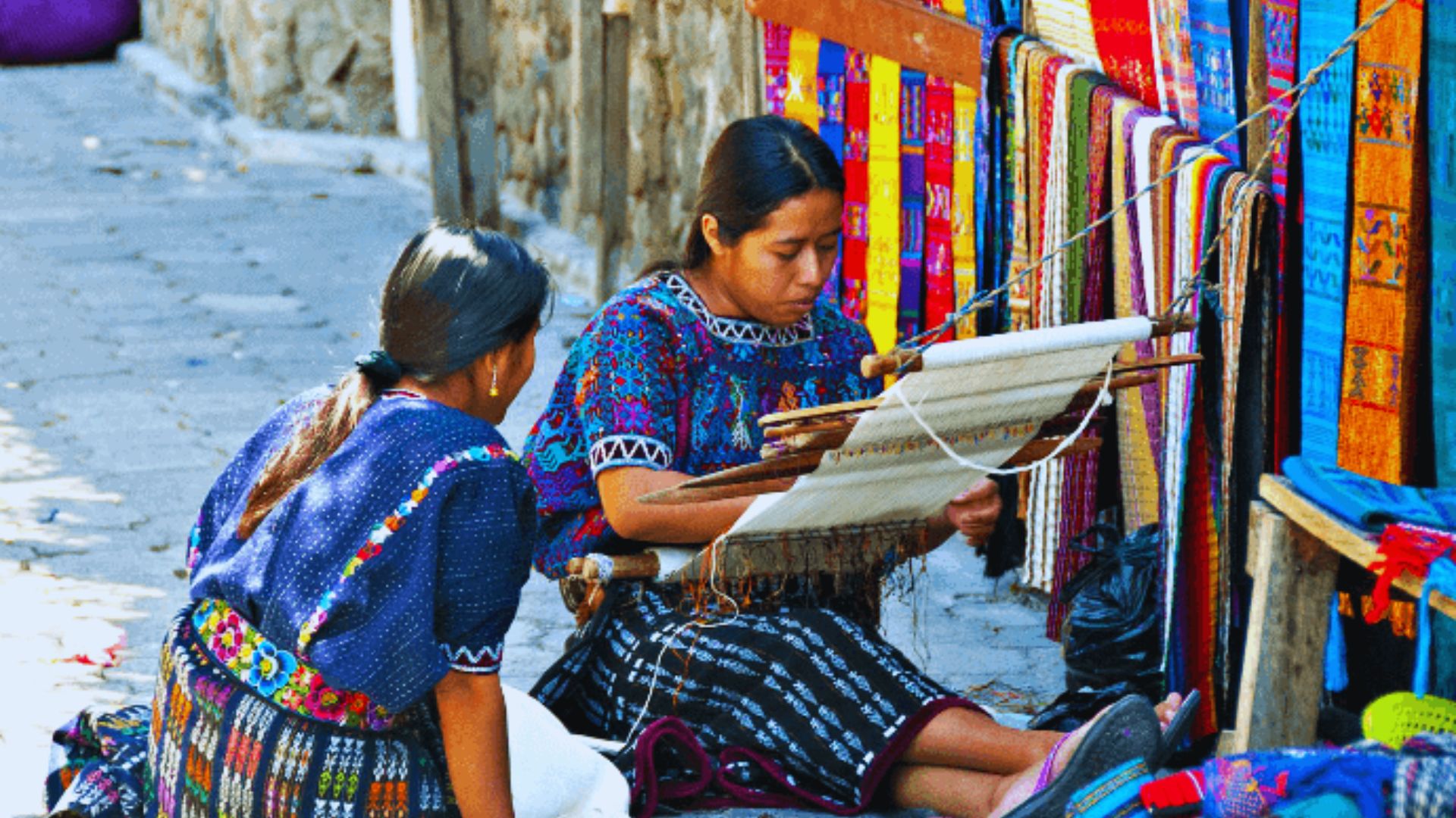Two women in traditional attire sit on the ground surrounded by vibrant textiles, showcasing cultural heritage and craftsmanship.