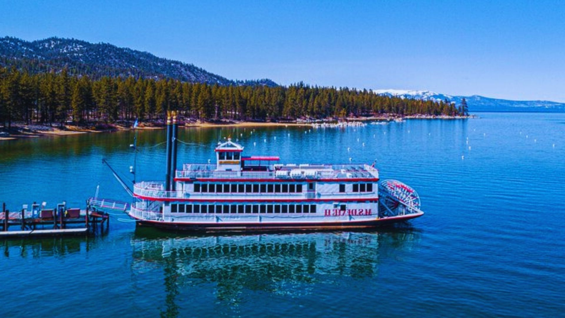A riverboat is docked at a lakeside dock, surrounded by calm waters and greenery.