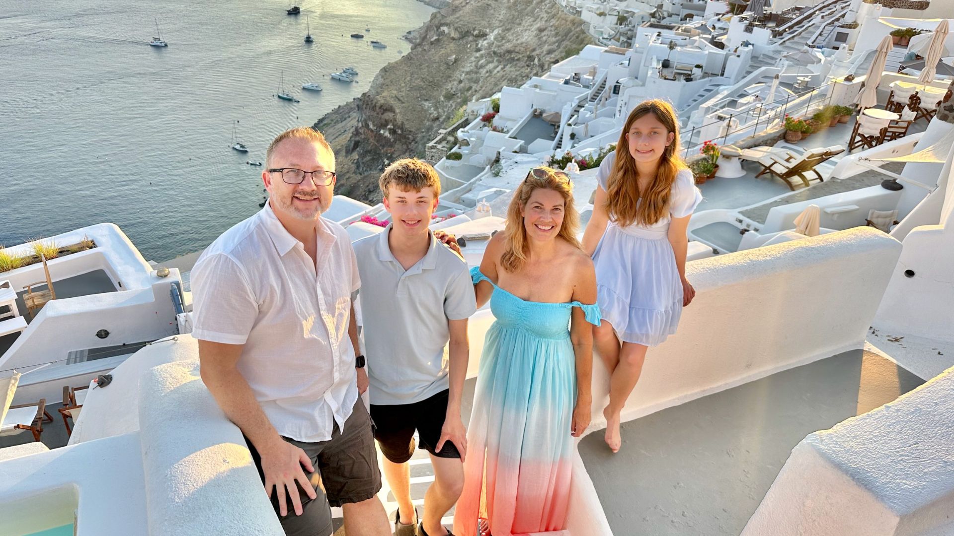A family of four smiles on a sunlit terrace overlooking the sea in Santorini. White buildings and the calm ocean create a serene backdrop.