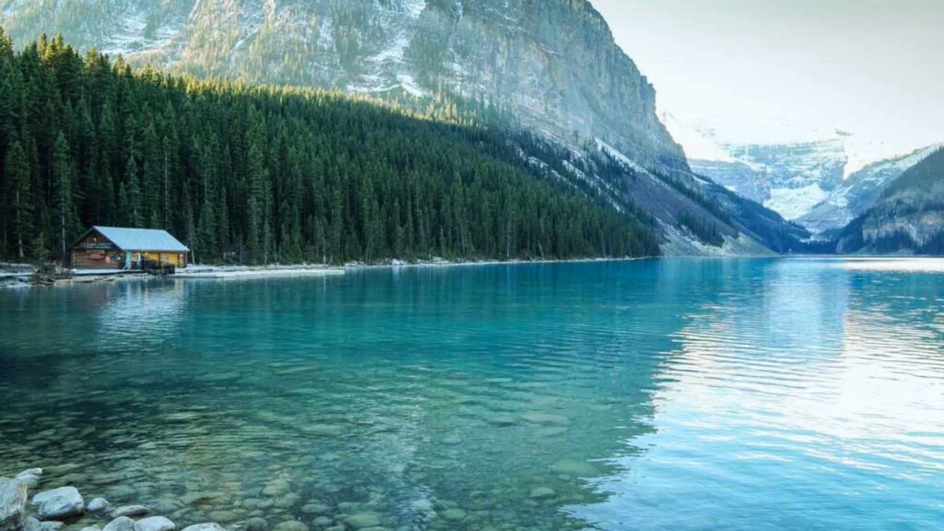 A serene turquoise lake reflects surrounding snow-capped mountains and dense pine forest. A small wooden cabin sits on the left shore, suggesting tranquility.