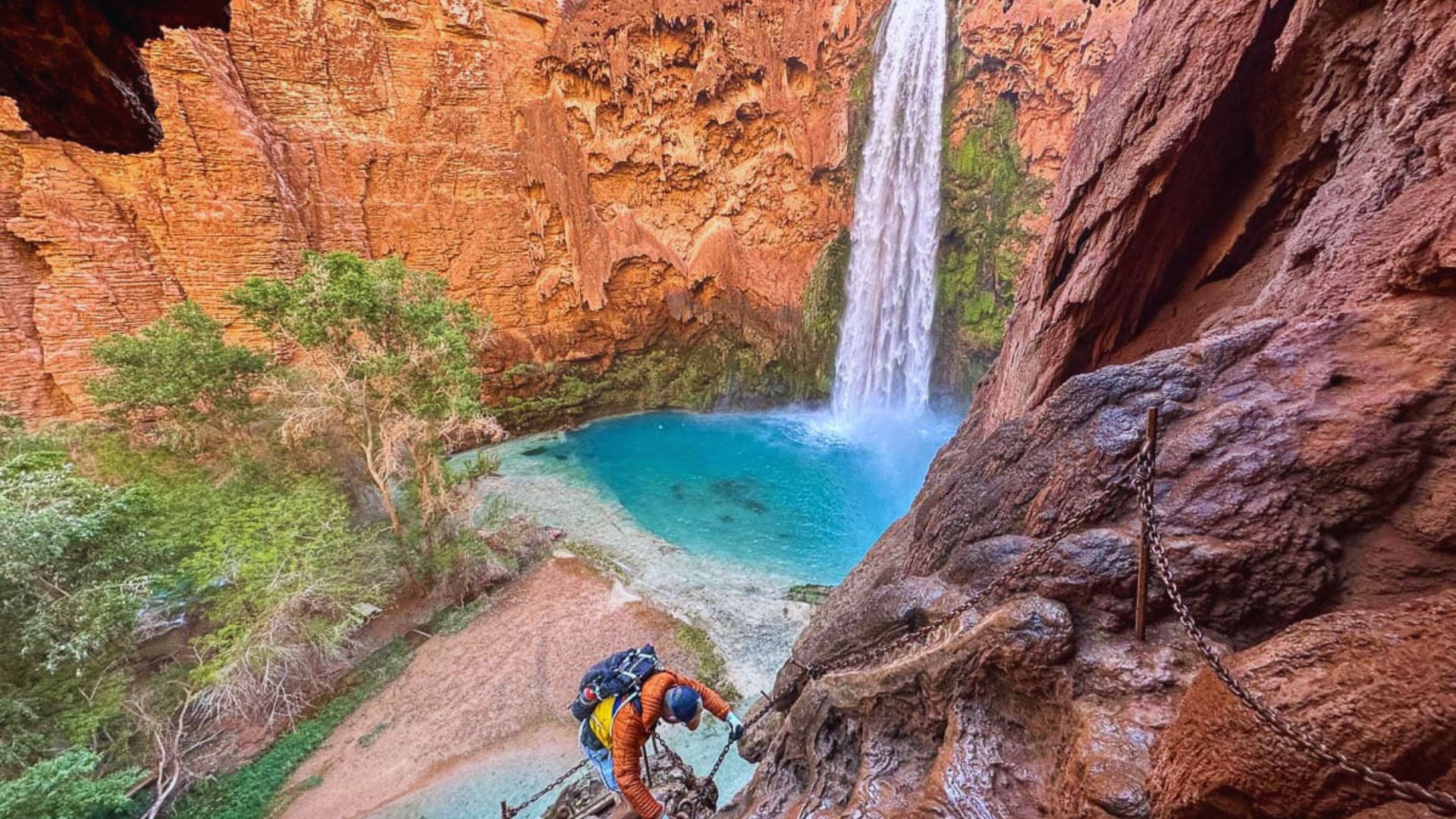 A hiker in an orange jacket climbs a rocky trail with chains beside a vibrant blue waterfall and pool, surrounded by steep red canyon walls and greenery.