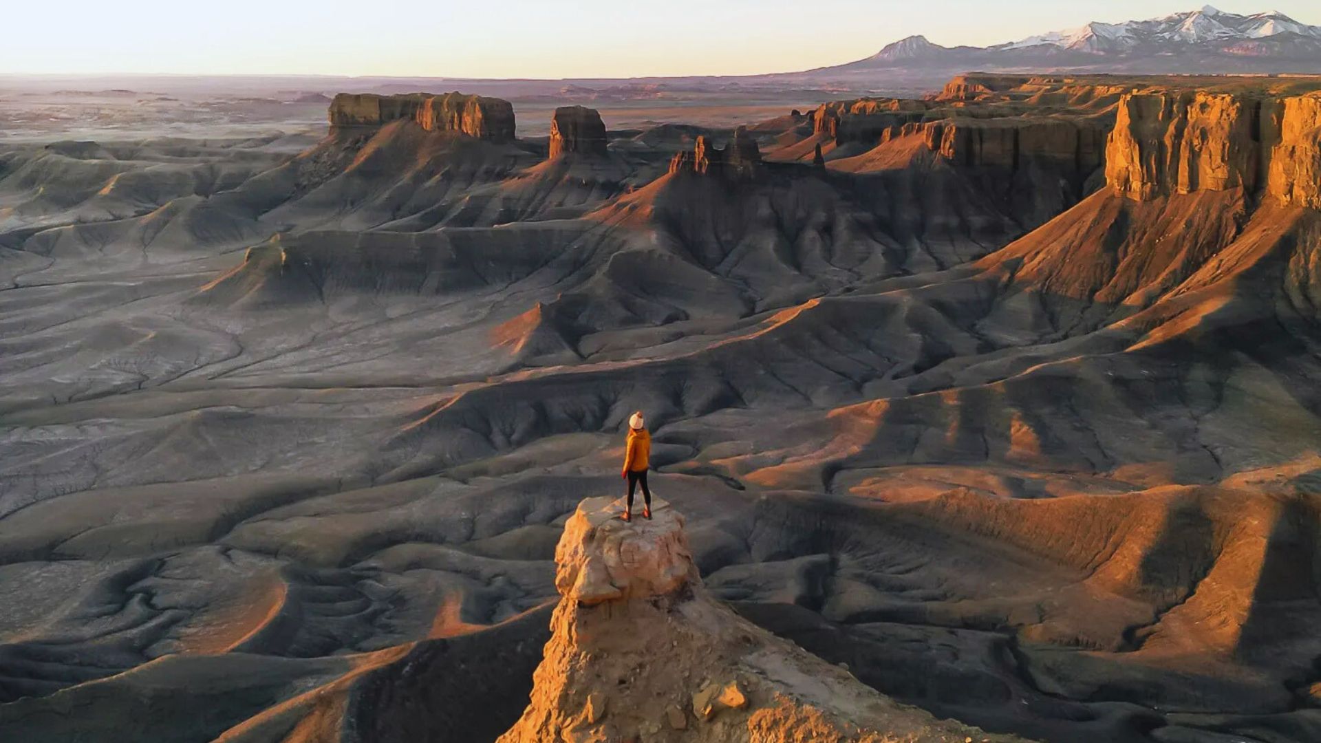 A person in an orange jacket stands on a rocky peak overlooking a vast, rugged desert landscape at sunset. The scene conveys solitude and awe.