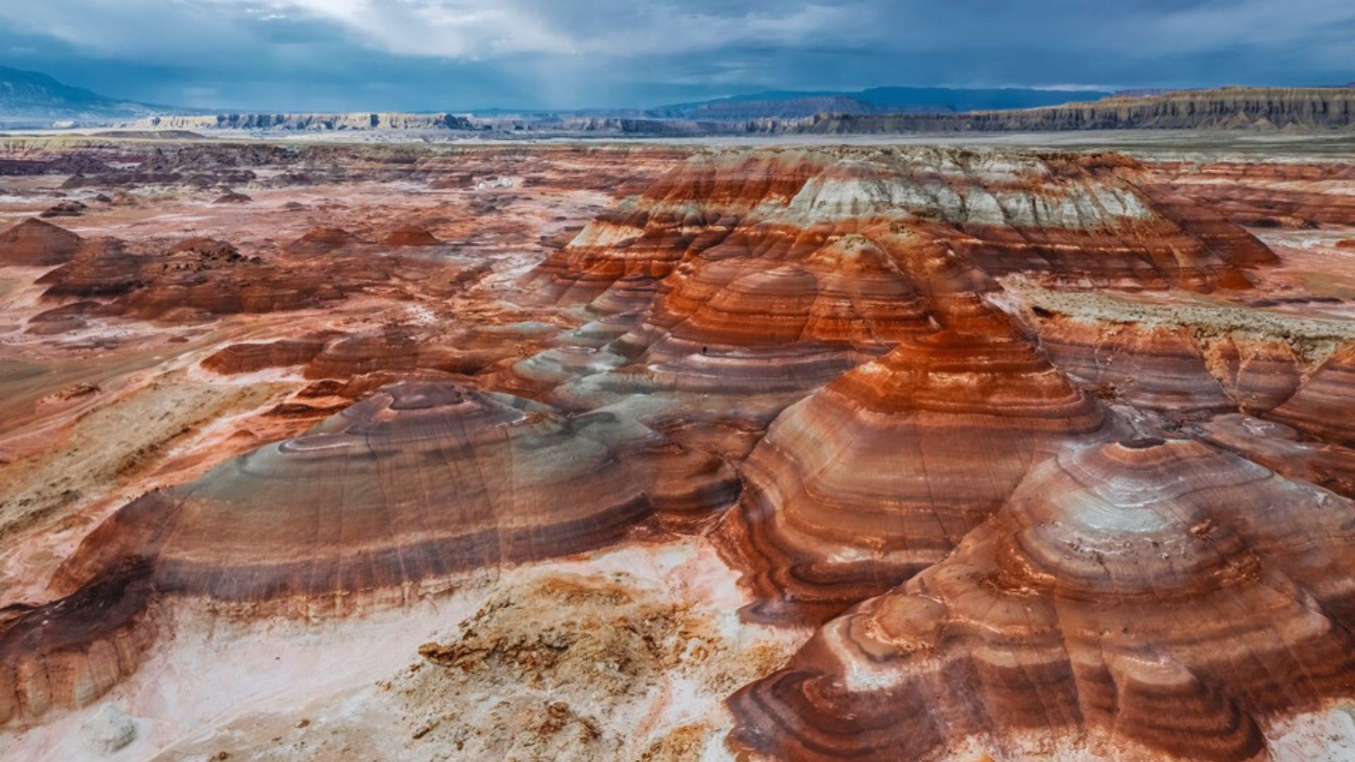 Vast desert landscape with layered rock formations in shades of red, orange, and white. The sky is overcast, creating a dramatic and serene atmosphere.