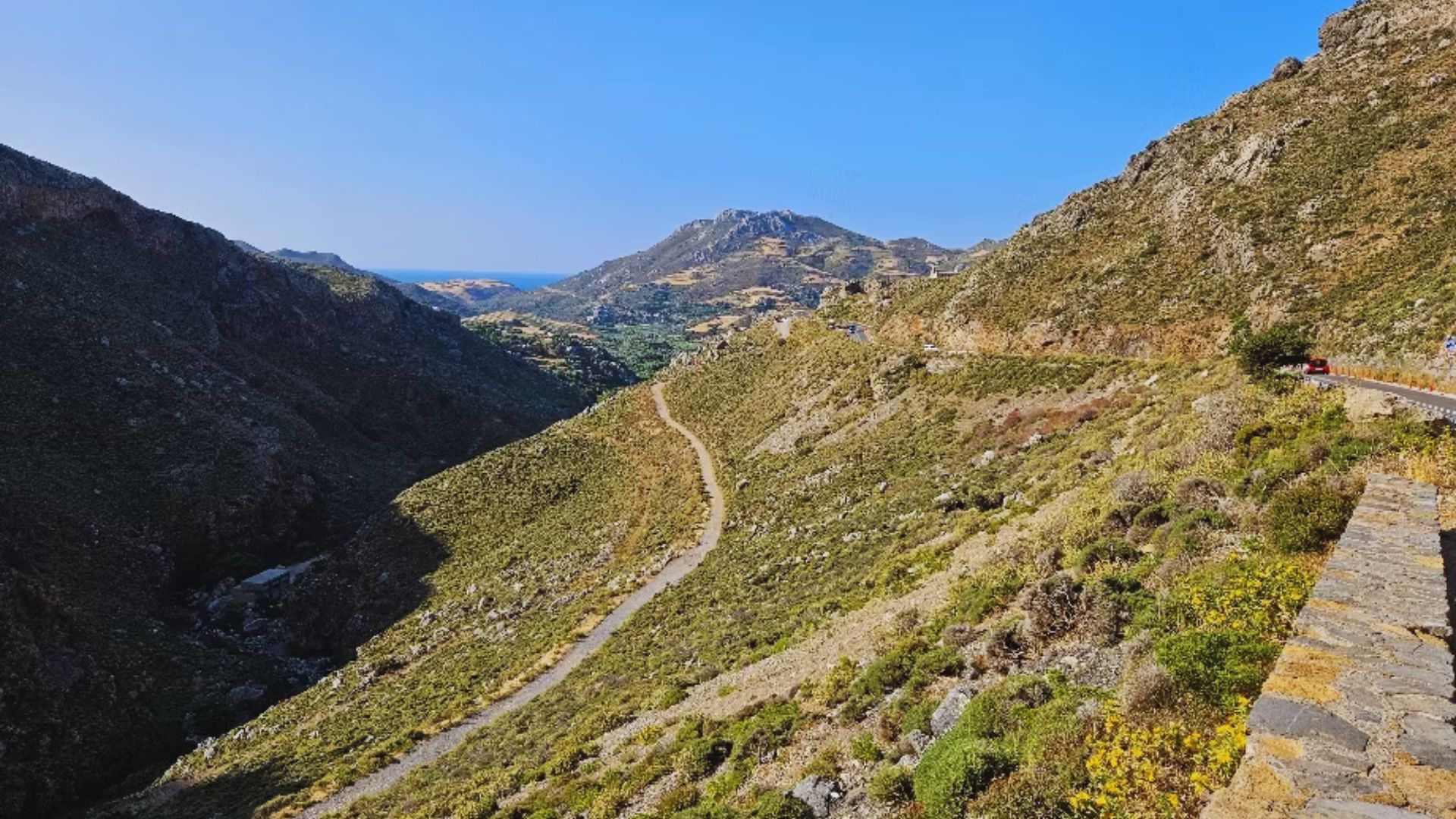 A winding road leading up a mountain, surrounded by lush greenery and rocky terrain under a clear blue sky.