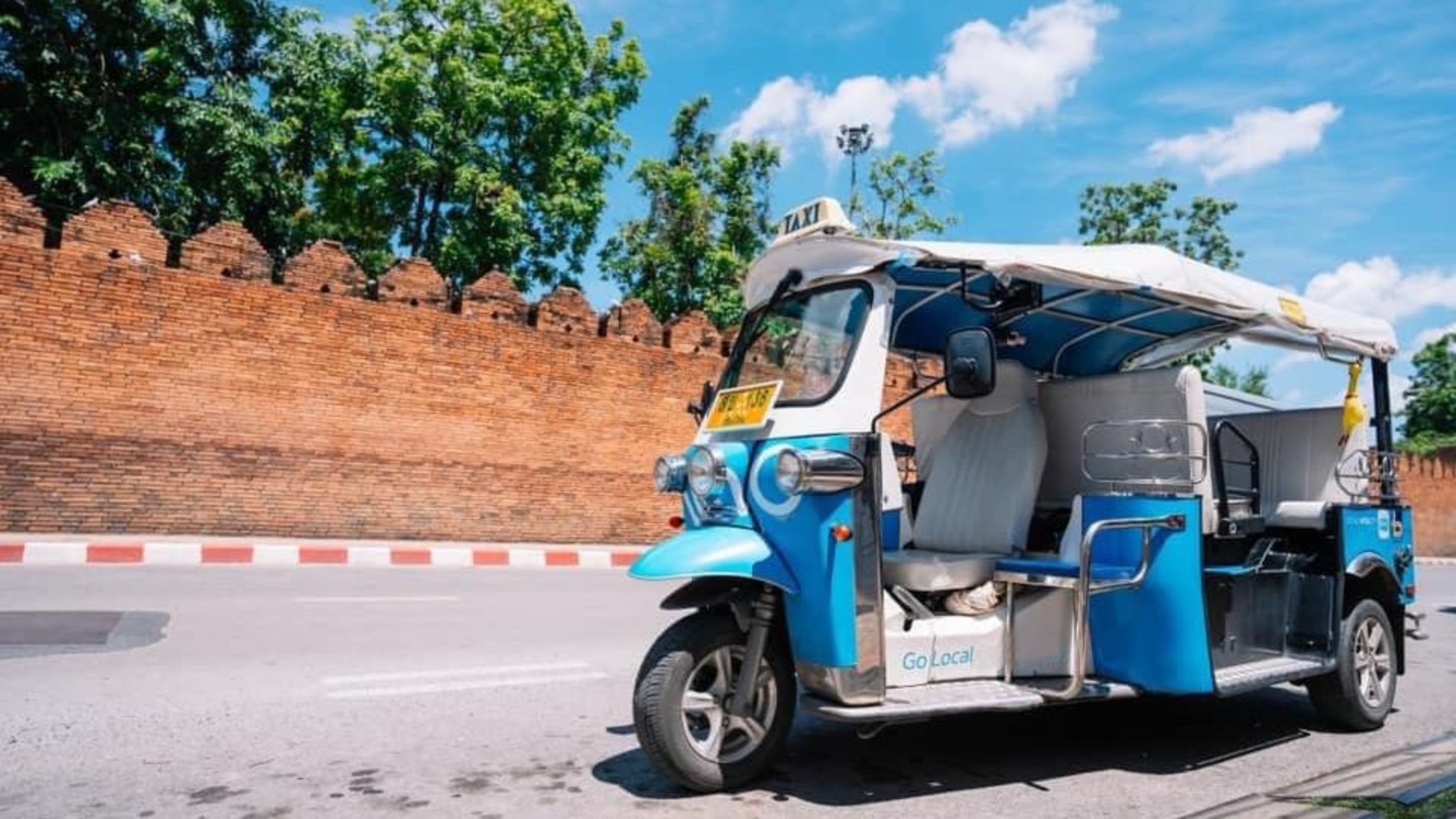 A blue and white tuk tuk parked on the roadside, surrounded by greenery and a clear blue sky.
