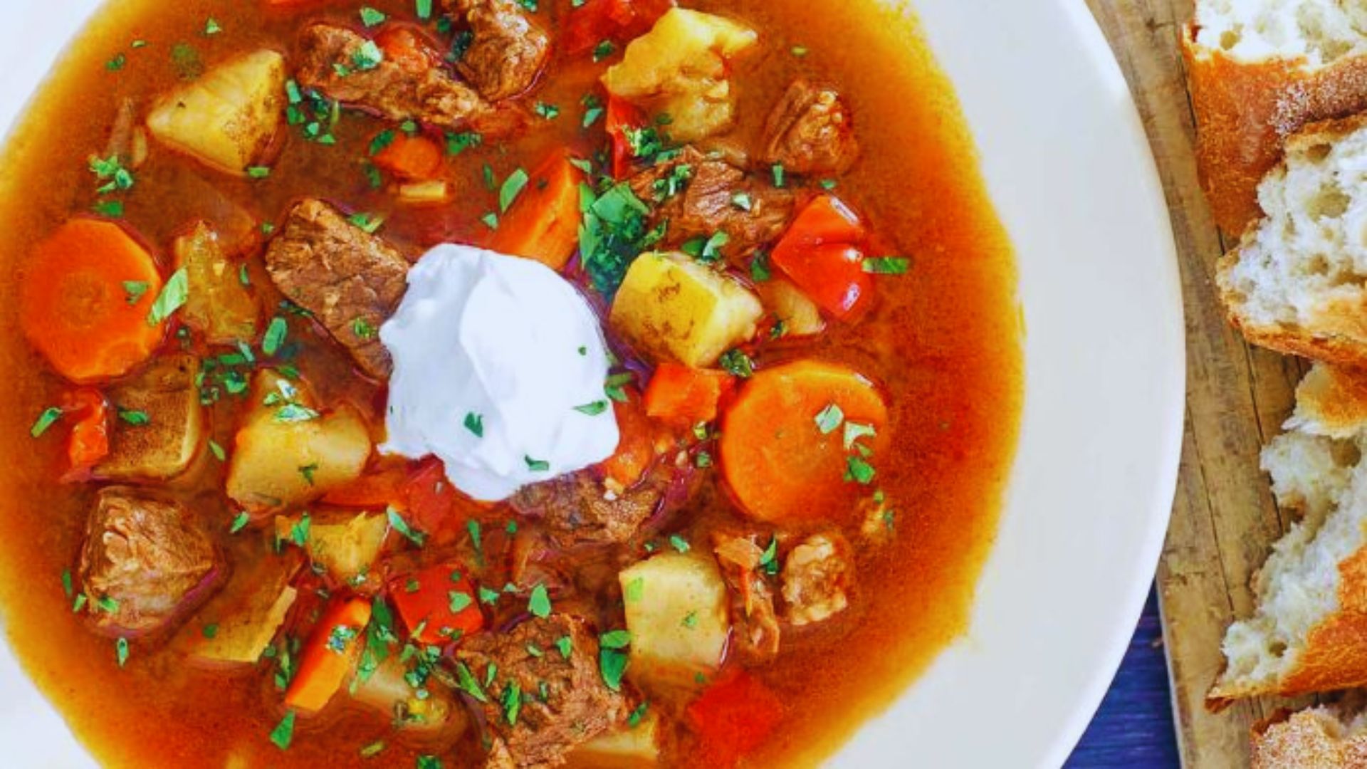 A bowl of hearty beef stew served with a side of bread and a dollop of sour cream.