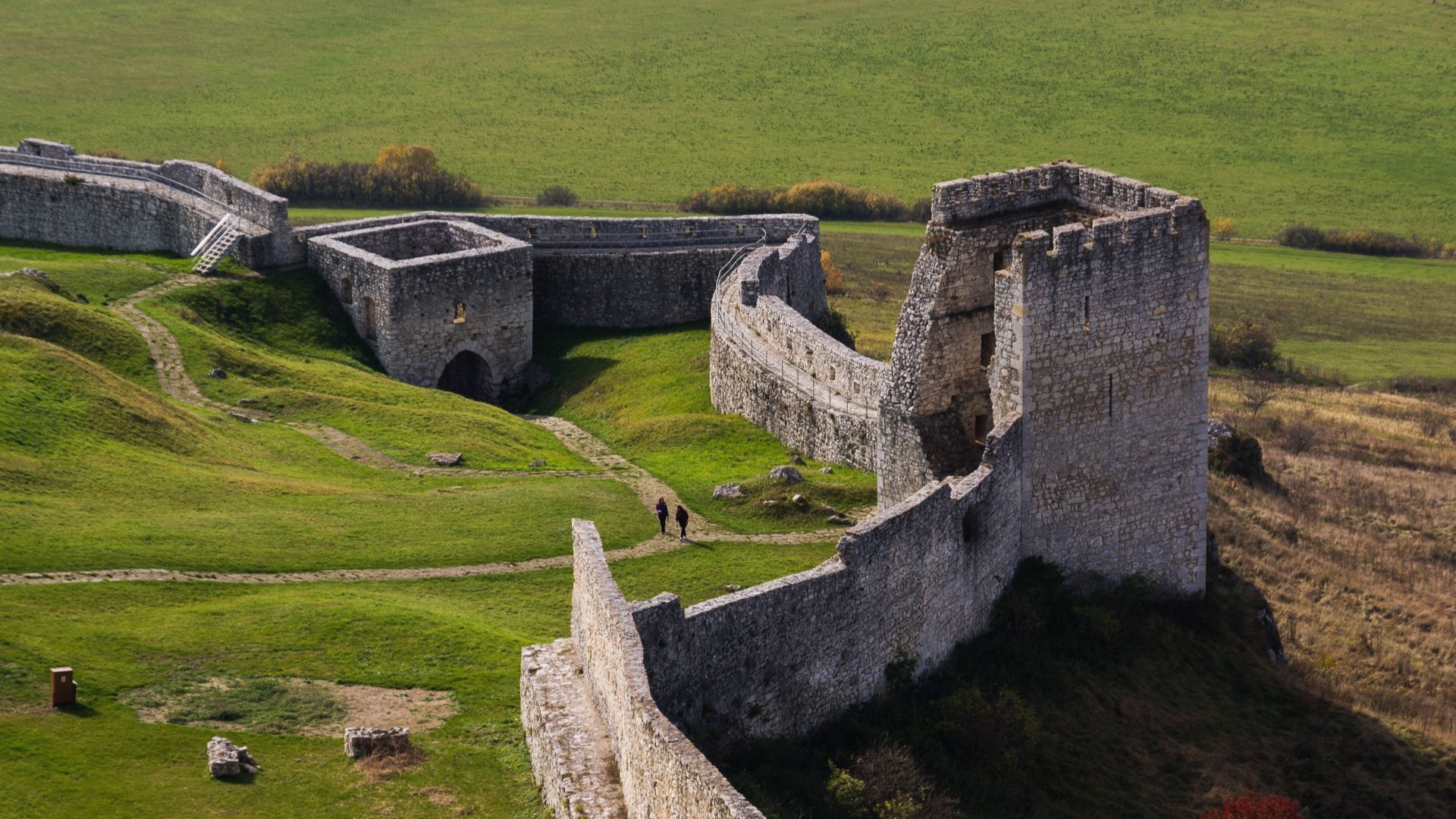 Ancient stone fortress with crumbling walls on a grassy hilltop. Two small figures walk along a path, surrounded by open, expansive countryside.